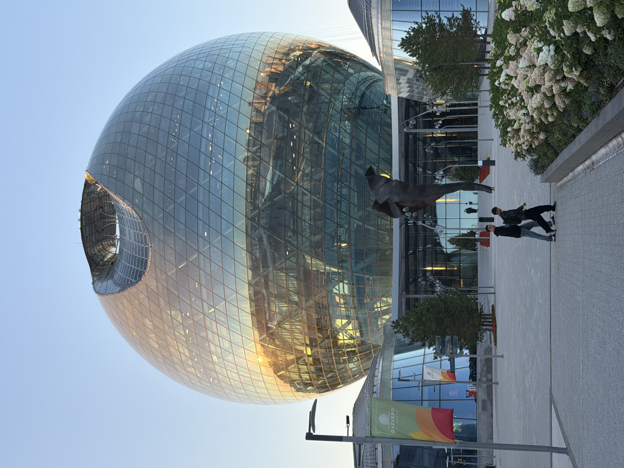 This is a medium-shot, eye-level photograph of the Palace of Peace and Reconciliation in Astana, Kazakhstan, also known as the Pyramid. The building is a prominent spherical glass structure with a geometric pattern of glass panels and a large opening at the top. The golden sunlight reflects off the glass, illuminating the building's surface. In front of the building, there is a modern plaza paved with light-colored stones. Two young men are walking across the plaza from left to right. One is wearing a black hoodie and blue jeans, and the other is wearing a black jacket and black pants. To the right of the plaza, there is a flower bed with blooming hydrangeas, displaying a mix of white and pink flowers. The sky is clear and blue, with the sun setting or rising, casting a warm glow on the scene. A tall streetlamp stands on the left side of the image, near a flag with "QAZEXPO" written on it.