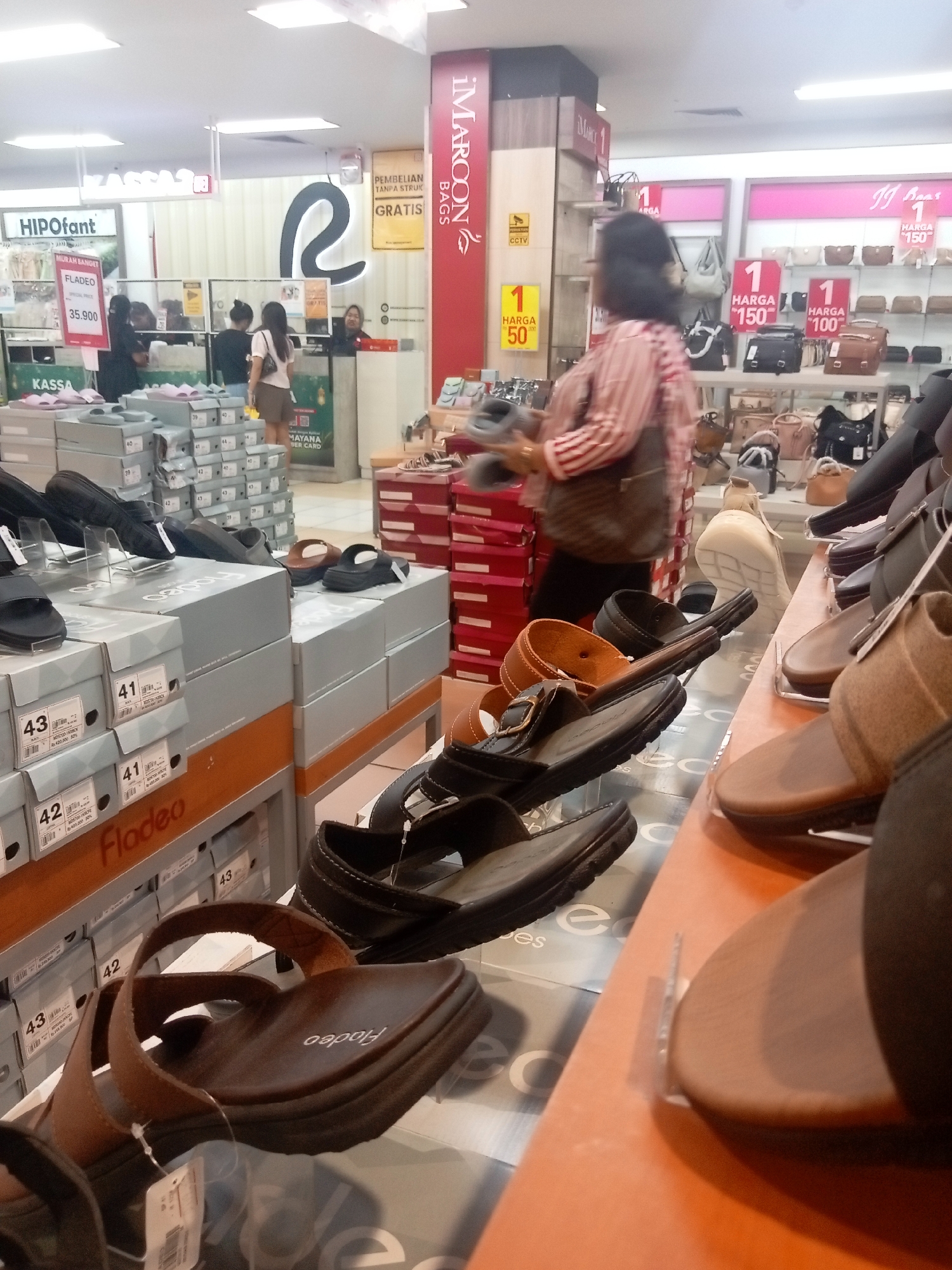 This image captures an interior scene within a retail store, likely a shoe or accessories shop, possibly located in Batam, Indonesia, given the presence of Indonesian currency (Rp) and common retail signage styles. The foreground and midground are dominated by displays of footwear and shoe boxes. Specifically, numerous shoe boxes are stacked, some with labels indicating sizes like 41, 42, and 43, and brand names such as "Fladeo." Several pairs of sandals and shoes are also displayed on shelves and counter-like surfaces. One prominent item in the mid-right foreground is a pair of brown sandals with the "Fladeo" logo visible on the sole.
In the background, a woman with dark hair is seen walking away from the camera, wearing a striped shirt and carrying a bag. To her left, two other women are standing near a counter area. Further back, a cashier is visible behind a counter, facing the camera. The store environment appears brightly lit with fluorescent ceiling lights. Signage in the background includes "HIPOfant," "MURAH BANGET," and other promotional text in Indonesian. A vertical red sign with "IMARCON BAGS" is also visible. The overall impression is of a busy retail environment with products arranged for sale.