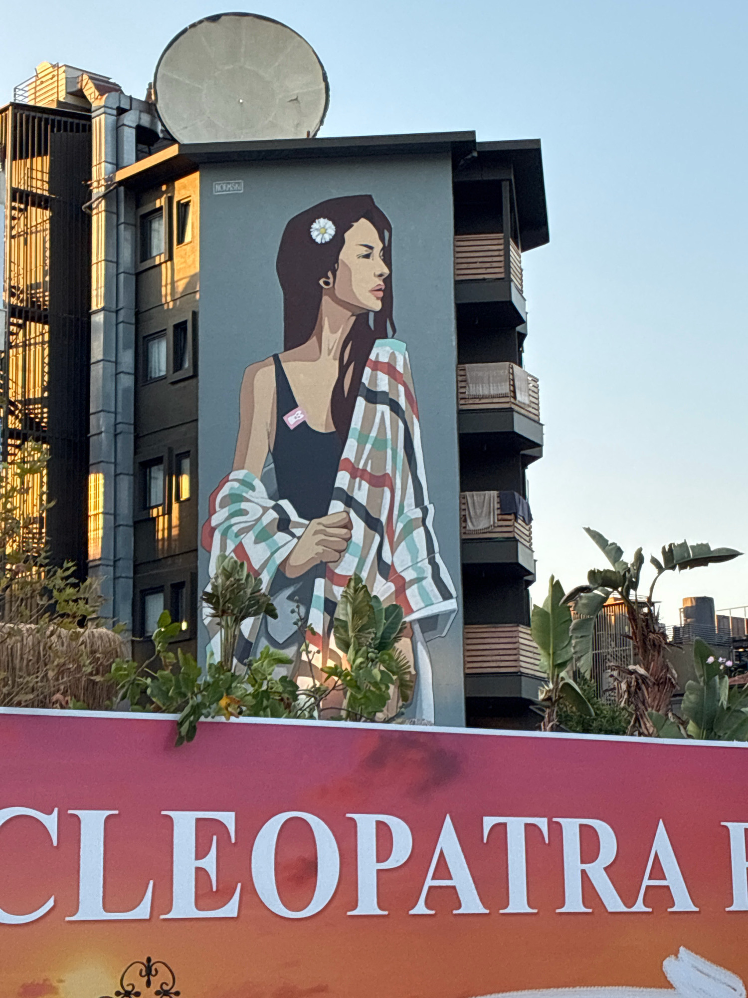 This is a photograph of a building in Alanya, Turkey, featuring a large mural of a woman. The mural covers a significant portion of the building's facade and depicts a woman with dark hair, wearing a black top and holding a colorful striped blanket. She has a white flower in her hair and a small pink tag on her top. The building itself is multi-story with balconies, some of which have laundry hanging on them. In the foreground, a large red banner with white lettering is partially visible, displaying the word "CLEOPATRA". The scene is illuminated by what appears to be morning or late afternoon sunlight, casting warm tones on the building and the surrounding foliage. The sky is clear and blue.