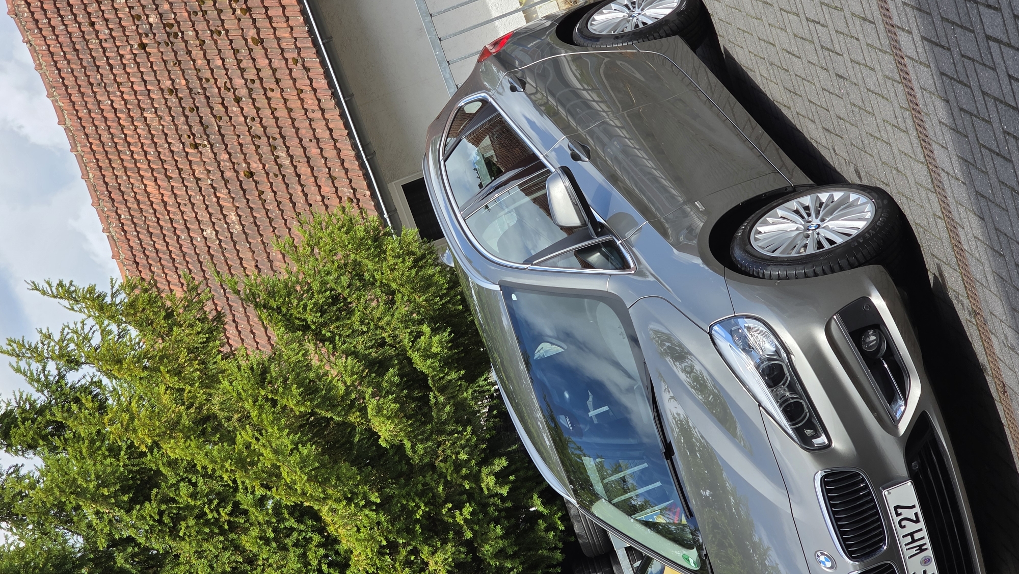 A grey BMW hatchback is parked on a brick-paved driveway in front of a building with a red tile roof and a large green tree. The car is angled towards the viewer, showing its front and side. The license plate reads "F-WH 27". The sky is partly cloudy. There are no people visible in the image.