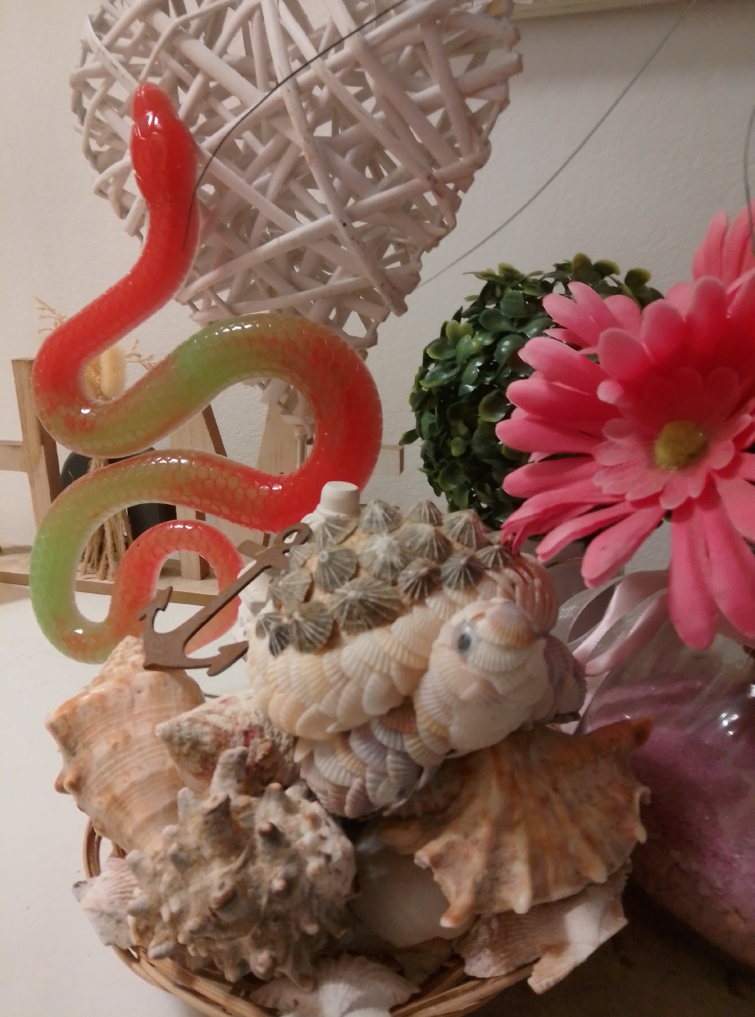 The image shows a decorative arrangement of shells, a red and green snake figurine, and a pink flower. In the background, a white heart-shaped wicker object and a green artificial plant are visible. The arrangement is placed on a white surface, possibly a table or shelf. The objects appear to be part of a home decor, likely in a domestic setting. There is no visible indication of the city of Wedel, Germany in the image.