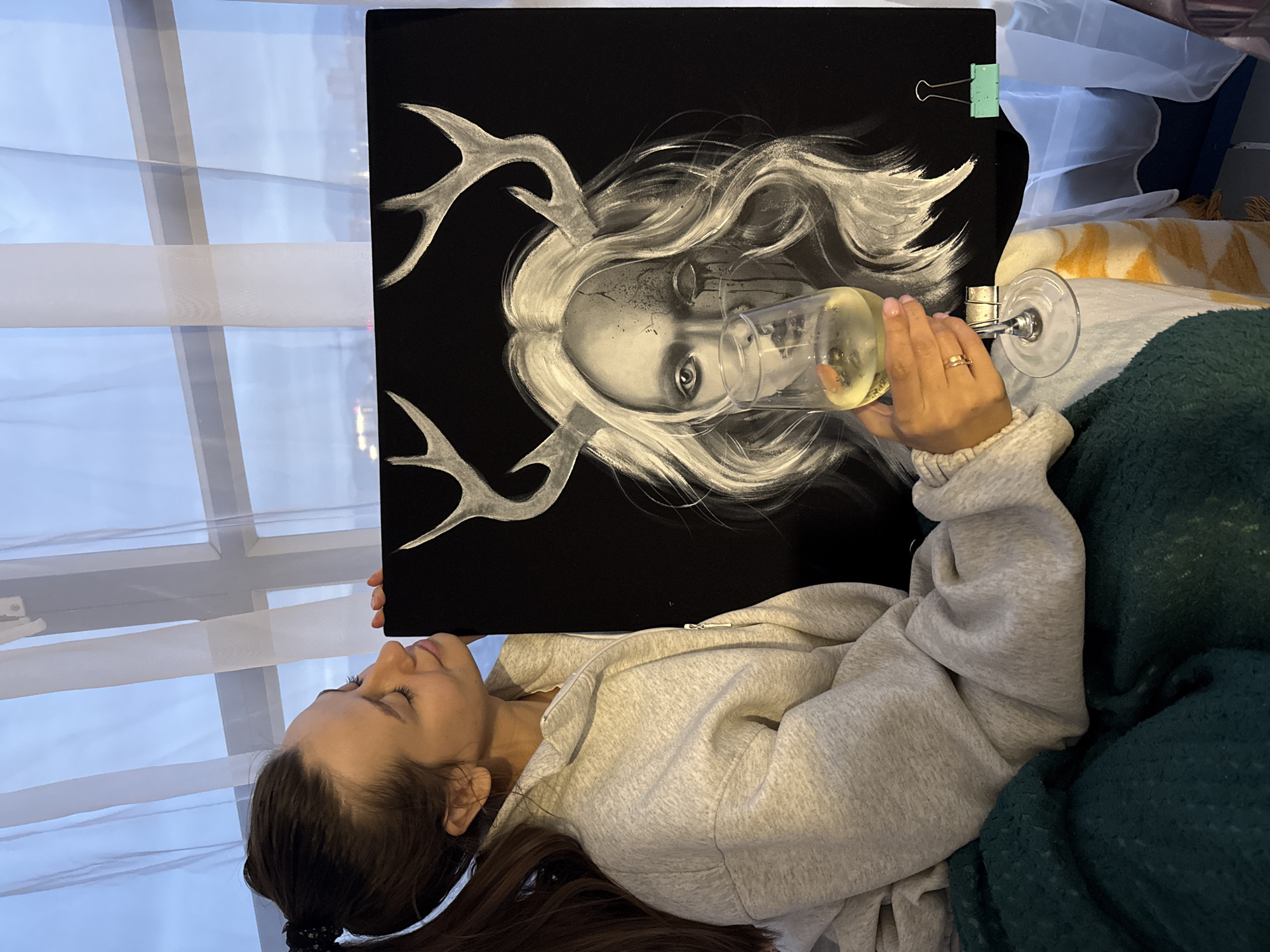 A person lies on a bed, holding a black canvas in front of them. The canvas depicts a stylized black and white drawing of a face with antlers. A wine glass filled with a yellow liquid is held in front of the face on the canvas. The person wears a light gray zip-up hoodie and a ring on their left ring finger. Behind the canvas is a window with sheer curtains. A yellow and white patterned pillow is visible behind the bed. A dark green blanket is also visible near the person. The image was taken in a room with a window, but there is no location given.