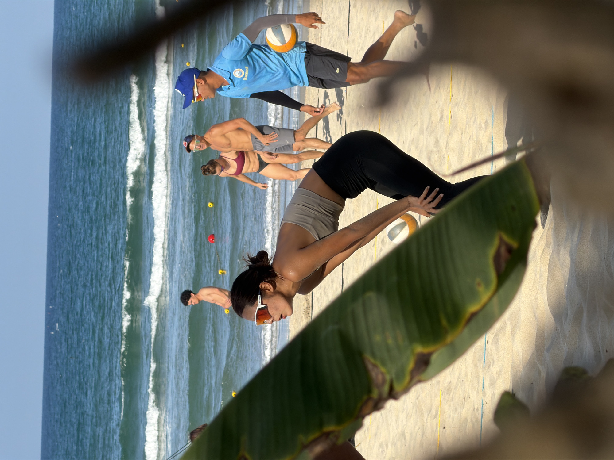 A low-angle shot from behind a large banana leaf with yellowed edges captures a beach scene, likely at Mỹ Khê Beach in Đà Nẵng, Vietnam, given the sandy beach and ocean. A woman in sunglasses, a cropped tan top, and black leggings is bent over in a stretch, her hands resting on a volleyball. Behind her, a volleyball game is in progress; a man in a blue shirt and cap is mid-jump, reaching to hit a volleyball. Further back, a couple and another man are also present on the beach near the water's edge. The ocean has visible waves, and the sky is pale blue. The foreground is partially obscured by shadows and other beach foliage.