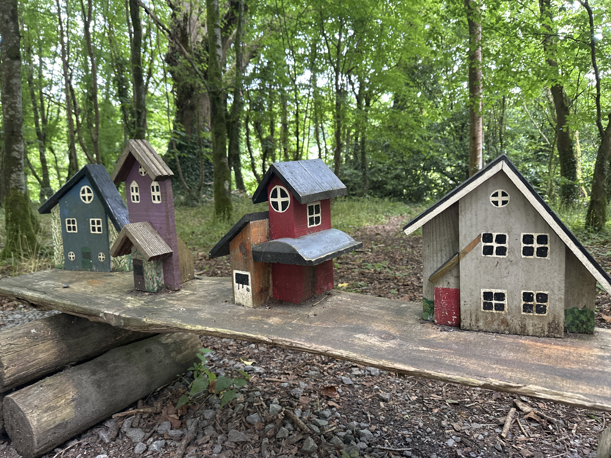 The image depicts a whimsical display of four miniature houses, crafted from wood and painted in various colors, arranged on a rustic, weathered wooden plank. The plank itself is elevated by two rough-hewn logs, creating a makeshift platform. The houses, from left to right, are painted dark green with a black roof, purple with a brown roof, red and orange with a grey roof, and a neutral grey with a black roof. Each house features miniature windows and doors, some with decorative elements like a cross shape within a circular window. The setting is a woodland environment, with tall trees forming a lush green canopy overhead. The ground beneath the platform is covered in small stones and fallen leaves. The scene appears to be located in the townland of Ballyfintan, in the parish of Castlecomer, County Kilkenny, Ireland. There are no people or animals visible in the image, and no actions or interactions are taking place.