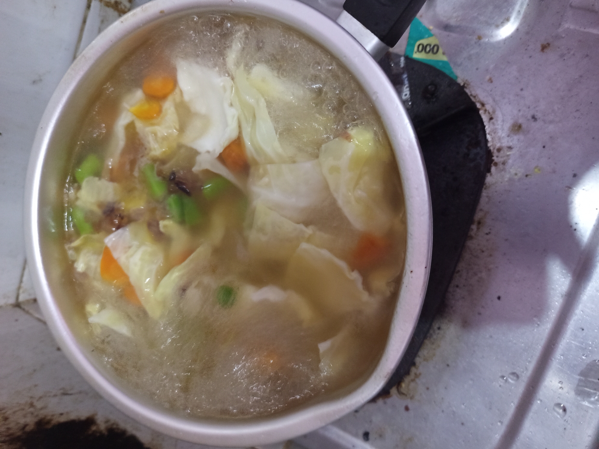 A close-up, overhead shot shows a pot of soup actively boiling on a stovetop. The soup contains visible pieces of cabbage, carrots, and what appear to be green peas and some darker, possibly meat ingredients. The pot is silver with black handles and is placed on a gray metal stovetop that shows signs of wear and some discoloration. The background is out of focus but suggests a tiled wall with some dark staining. A small, green and yellow sticker with the number "000" is visible on the stovetop to the right of the pot. The image appears to have been taken in a domestic setting, possibly a kitchen. The location in Tebel, Indonesia, is not visually discernible from the image content itself.