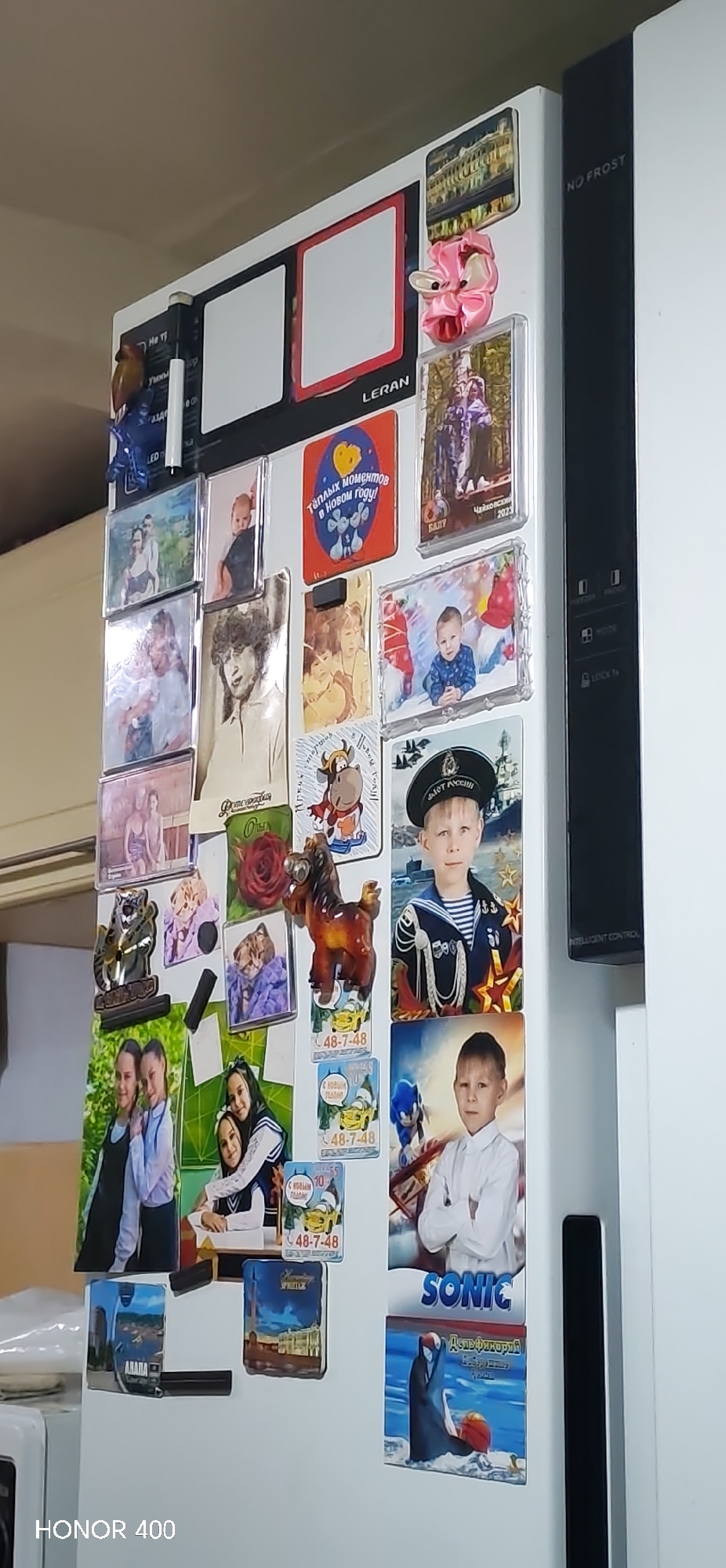 The image shows a white refrigerator covered in a variety of magnets and photographs. A prominent feature is a pair of framed white rectangles, with the brand name "LERAN" printed beside one. There are numerous photographs, including portraits of children and pictures of groups of people. Some of the photos appear to be school portraits or formal photographs. There is a magnet with the text "Тёплых моментов в Новом году!" A magnet of a cartoon cow is also visible. Several magnets relate to locations, including one for "Чайковский 2023." Magnets that mention, Anapa. Several magnets have Cyrillic text, while a "SONIC" magnet shows a dolphin playing with a ball. A horse statue appears to be attached to the refrigerator, presumably as a magnet. The refrigerator also has a "NO FROST" label and a control panel. The HONOR 400 phone is at the bottom left corner. The environment is somewhat domestic.
