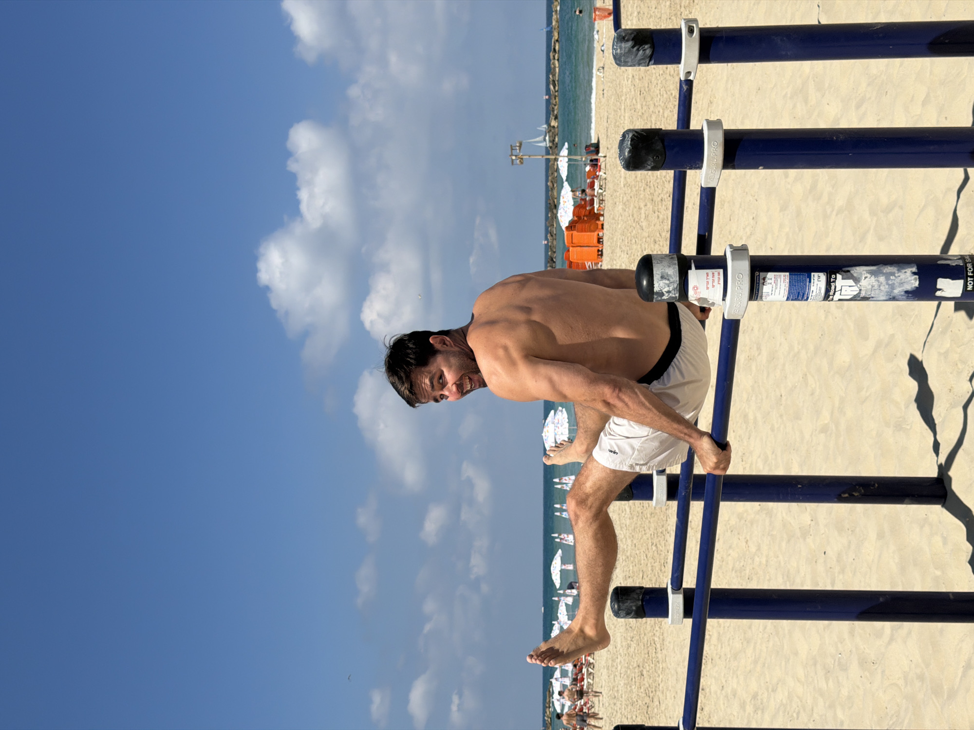 A shirtless man with dark hair and a beard is performing a calisthenics exercise on parallel bars at a beach in Tel Aviv, Israel. He is wearing light-colored shorts and is looking back at the camera with a smile. His legs are extended forward, and his arms are supporting his body on the bars. The background shows a sandy beach with beach umbrellas, lounge chairs, and people in the distance. The sea is visible behind the beach, with sailboats on the horizon under a blue sky with white clouds. The parallel bars are blue and appear to be made of metal. The overall scene depicts a person engaging in outdoor fitness in a popular public space.