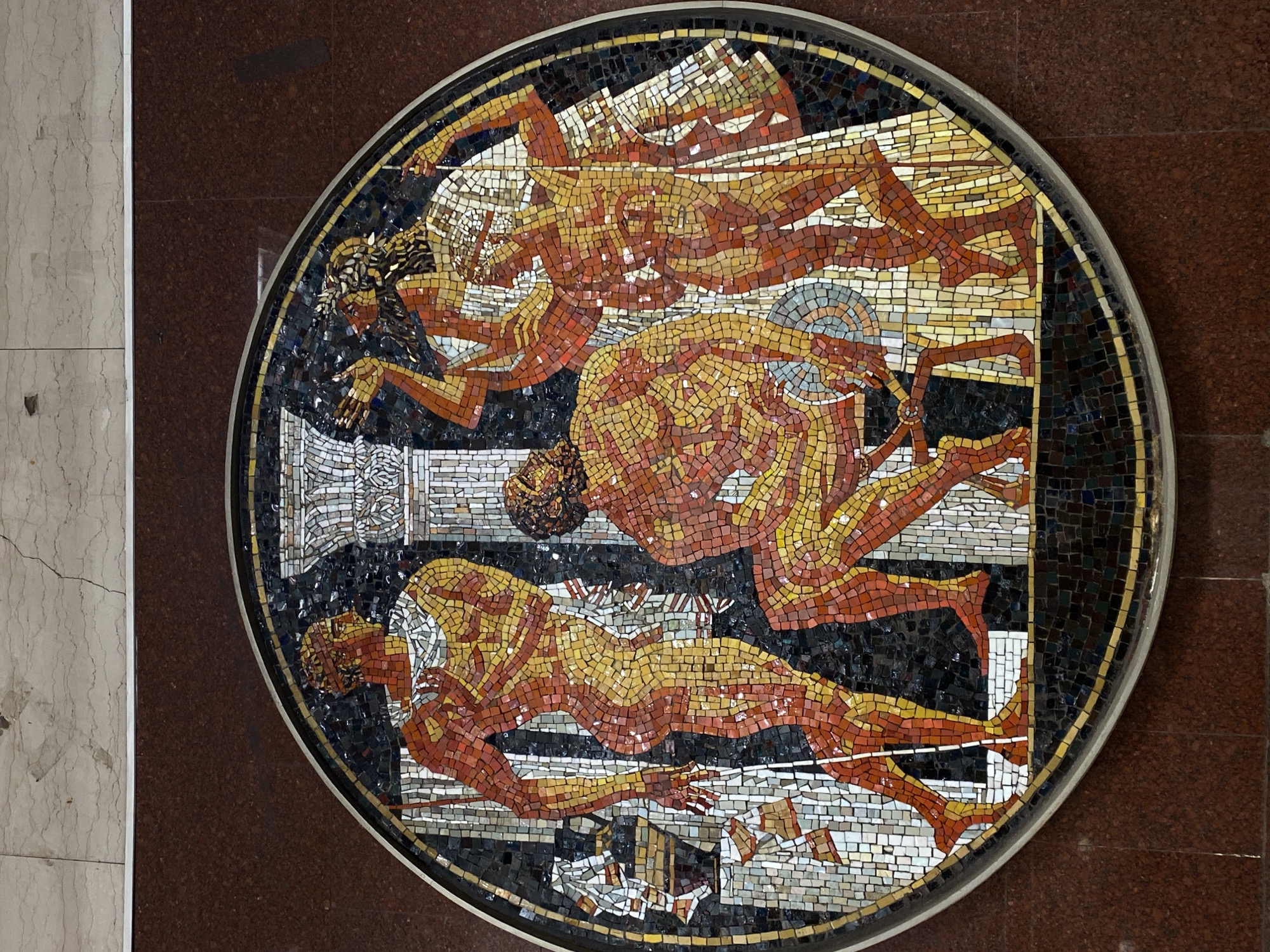 The image shows a circular mosaic depicting three figures in classical poses. The figures are rendered in shades of red, orange, and yellow mosaic tiles against a background of black and white tiles. The mosaic is set into a wall with brown and beige stone panels. The mosaic is located in the Saint Petersburg Metro, Russia, specifically in the Avtovo station. The mosaic depicts three figures and their associated actions: one figure is holding a column and appears to be falling, another is holding a round object, and the last one is laid out and wearing white clothing.