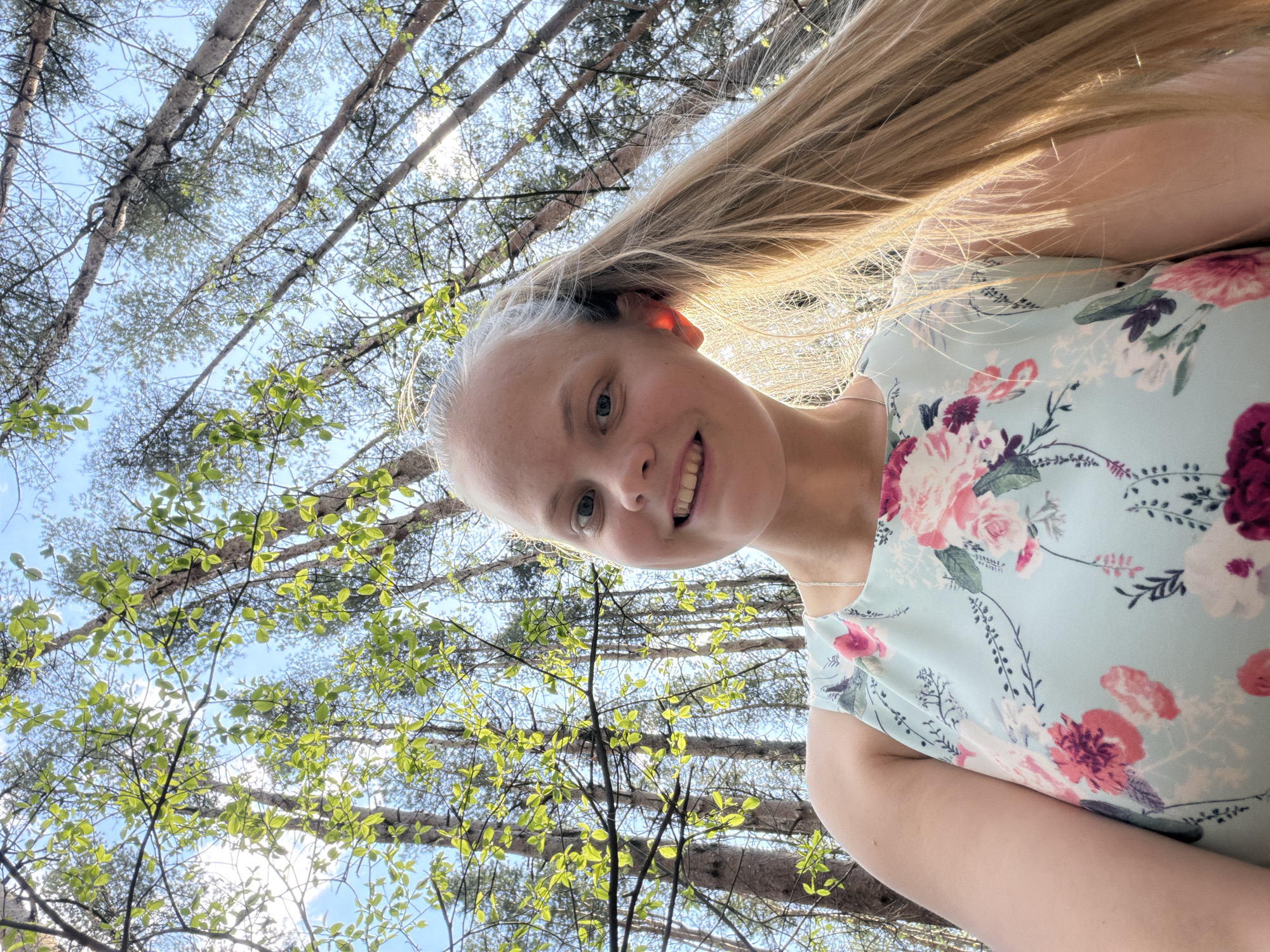 A woman with blonde hair and fair skin is smiling at the camera. She is wearing a light blue floral dress. The background consists of trees with green leaves against a blue sky with some white clouds. It is impossible to confirm this location is Izhevsk, Russia.