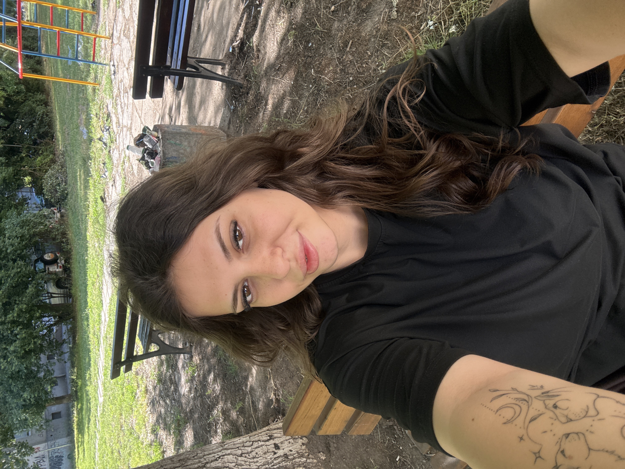 A young woman with long, dark brown, wavy hair and dark eyes is taking a selfie outdoors. She is wearing a black t-shirt and has a tattoo on her left arm. She is sitting on a wooden bench in a park-like setting. In the background, there is a trash can overflowing with debris, another park bench, trees, grass, a building with graffiti, and a playground structure. The scene appears to be in a public urban space, possibly a park or a courtyard. The overall lighting suggests it is daytime.