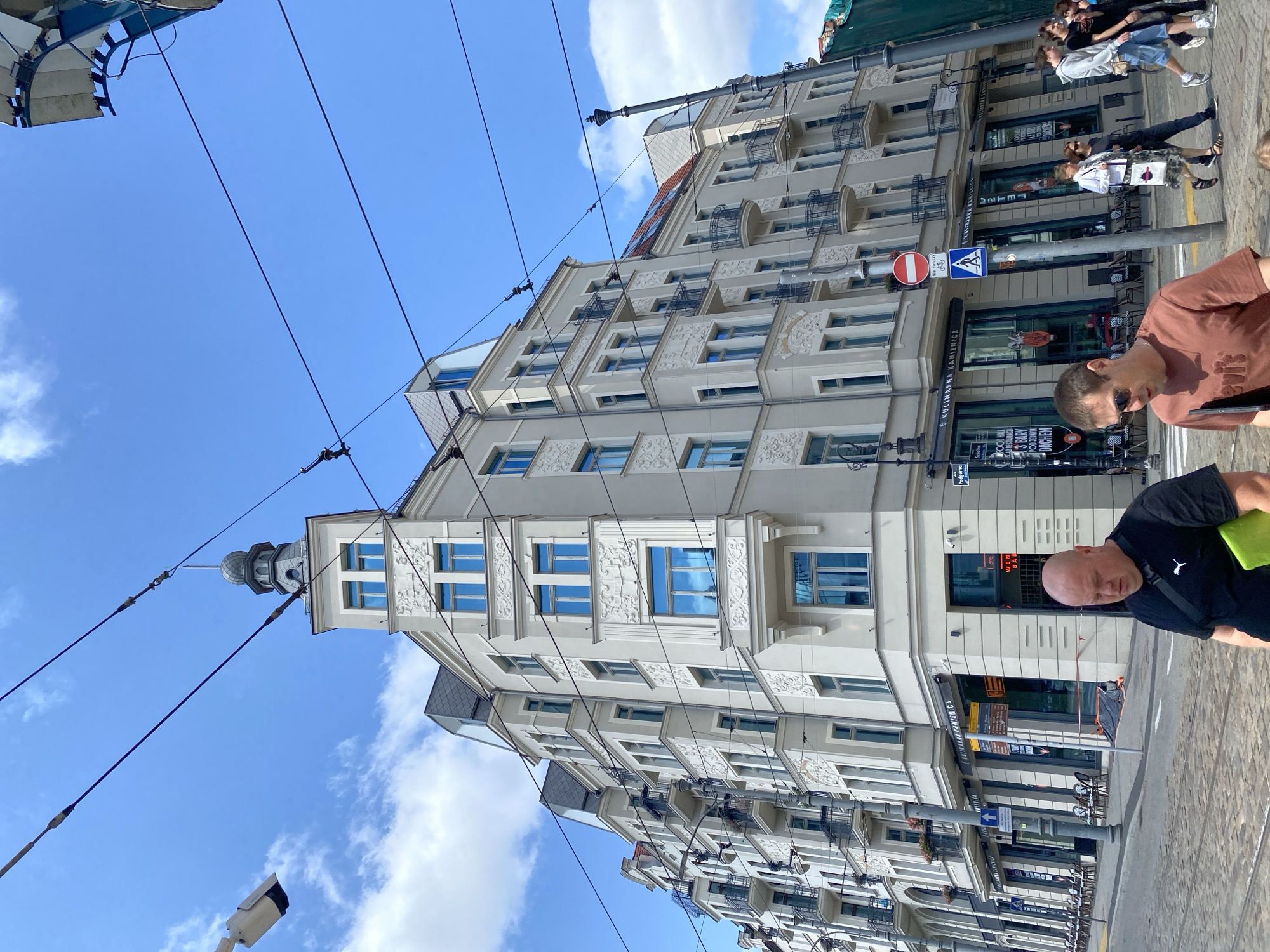 In Poznan, Poland, on a clear day with scattered clouds, a street-level view captures a multi-story building with ornate architectural details and blue-tinted windows. The building is situated at a corner, with the street in the foreground paved with cobblestones. Overhead, numerous power lines crisscross the sky, suggesting the presence of public transportation like trams.
Several people are walking on the sidewalk and crossing the street. In the immediate foreground, two men are visible. One, wearing a black t-shirt with a white logo, is walking away from the camera, carrying a bright green folder. The other man, in a brown t-shirt, is walking towards the camera. Further down the street, a group of individuals, including two women and several men, are walking together.
The building itself features a tower-like structure with a small dome and spire at the corner. The facade is light-colored, adorned with decorative reliefs around the windows and balconies. Street-level businesses are visible, with signage including "KUCHARNIA KAMIENICA". Outdoor seating with chairs and tables is arranged in front of one establishment. Traffic signs and streetlights are also present. The overall scene depicts an urban environment with ongoing pedestrian activity.