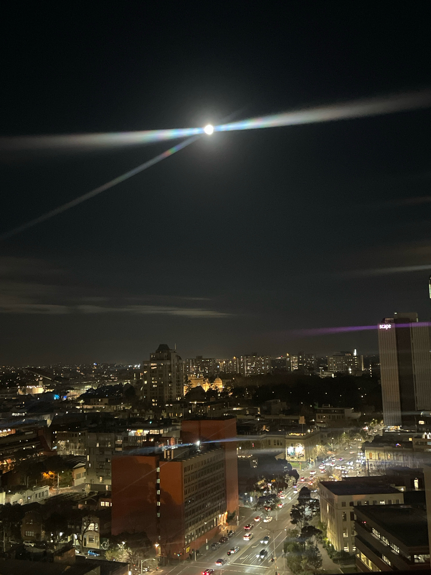 This is a nighttime aerial photograph of the city of Melbourne, Australia, taken from a high vantage point. The moon is visible in the upper portion of the frame, creating a bright lens flare with a prismatic effect. Below the moon, the cityscape is illuminated by streetlights and the lights of buildings.
In the foreground, a large, reddish-brown building with numerous illuminated windows dominates the lower left and center of the image. To the right of this building is another tall, modern building with a prominent "scape" logo illuminated in purple. The middle ground shows a street with a moderate amount of traffic, indicated by the headlights and taillights of cars. Various other buildings of different heights and architectural styles are visible throughout the cityscape, some with lit windows, others dark. Trees and patches of darkness suggest parks or less developed areas. The sky is dark, with some faint clouds visible, and the overall atmosphere is one of an active urban environment at night. No people are discernible in the image.
