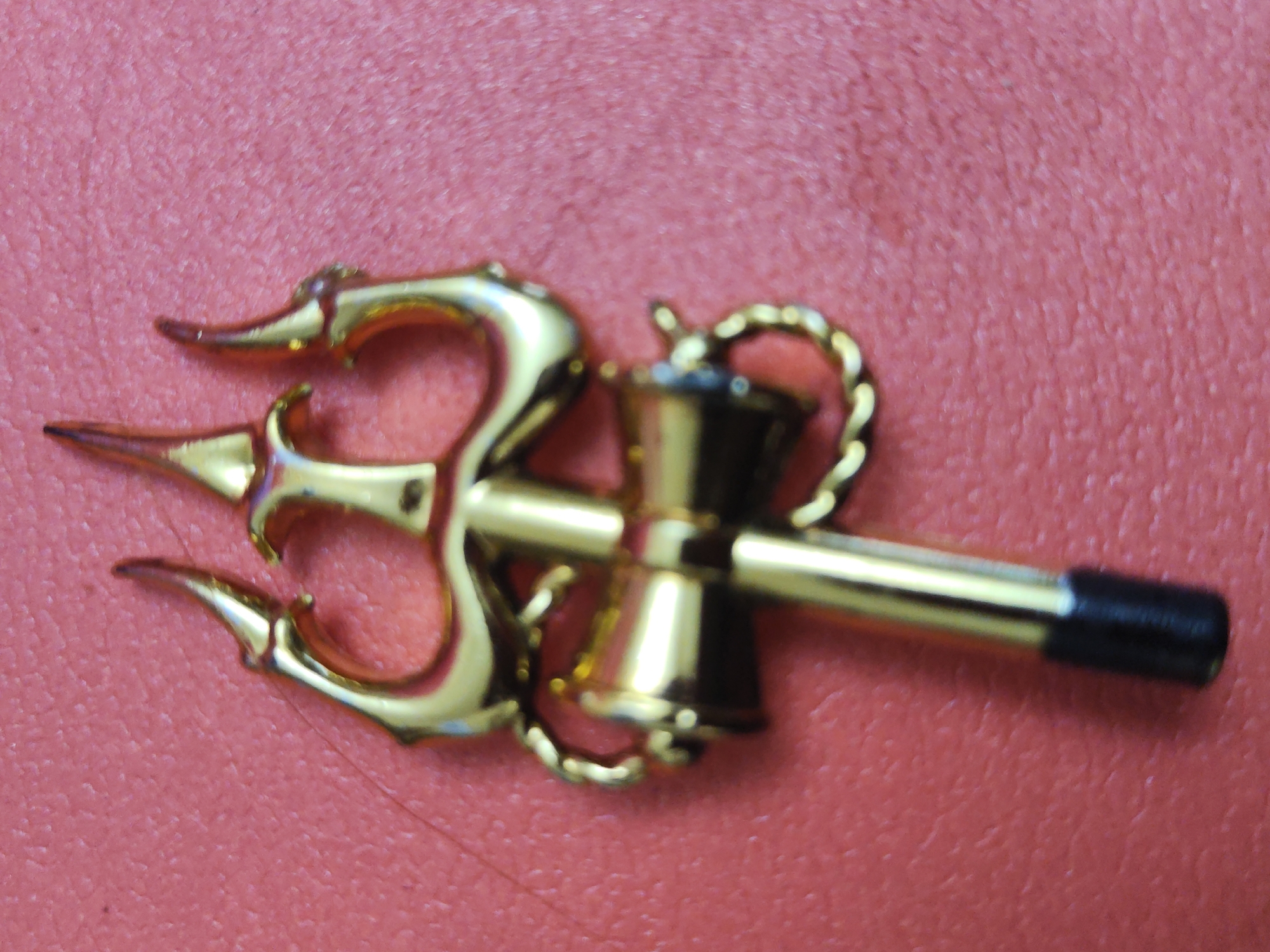 A gold-colored metal trident is positioned horizontally on a red textured surface. The trident has three prongs on the left side and a decorative element resembling a hammer attached to the right. The handle of the hammer extends to the right and is tipped with a black cap. The object is laying on a red textured surface.