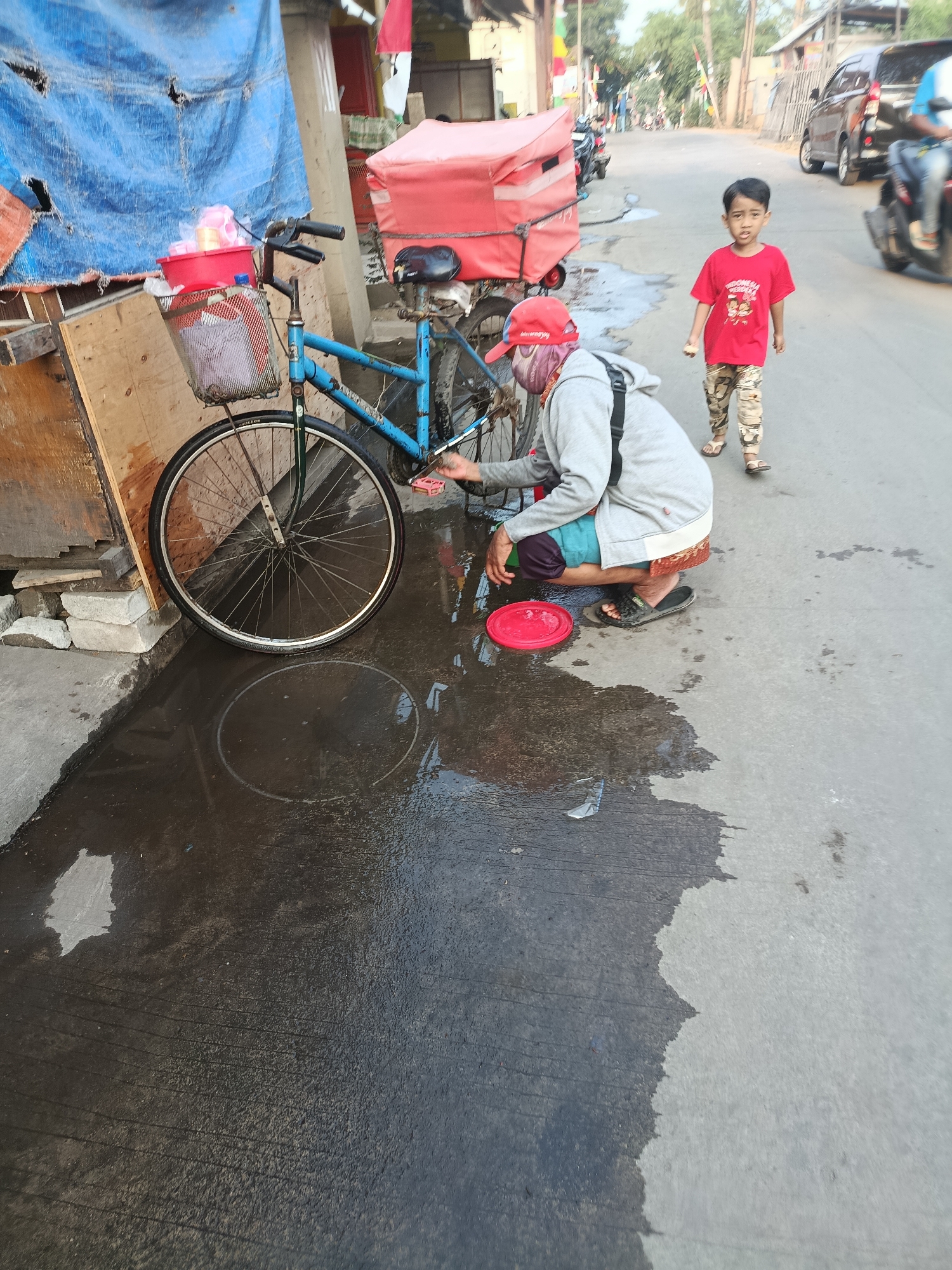 In Balaraja, Indonesia, a young boy wearing a red t-shirt and patterned pants walks on a wet street past a person hunched over a blue bicycle. The person, wearing a grey hoodie, a red cap, and a teal and brown garment, is cleaning the bicycle. A red plastic basin sits on the ground near the bicycle's front wheel. A large puddle reflects the bicycle's wheel. In the background, a red cooler box is visible, along with other vehicles and buildings lining the street. The scene appears to be a roadside or market area.