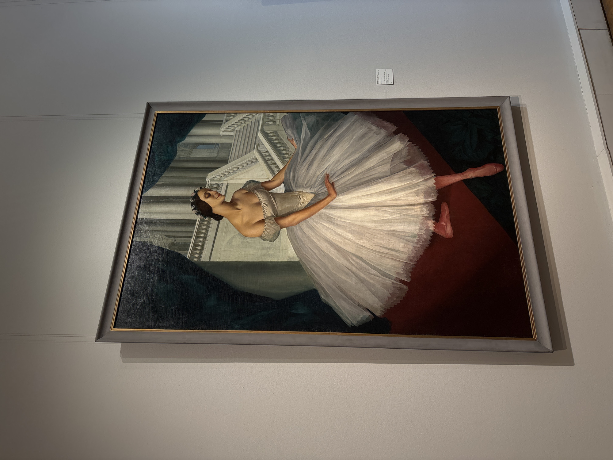 A framed painting hangs on a pale wall. The painting depicts a ballerina in a white tutu. She is surrounded by an architectural background and a red and dark green carpet. The painting is framed with a gray frame. There is a small sign above the painting. The location is likely in Moscow, Russia.