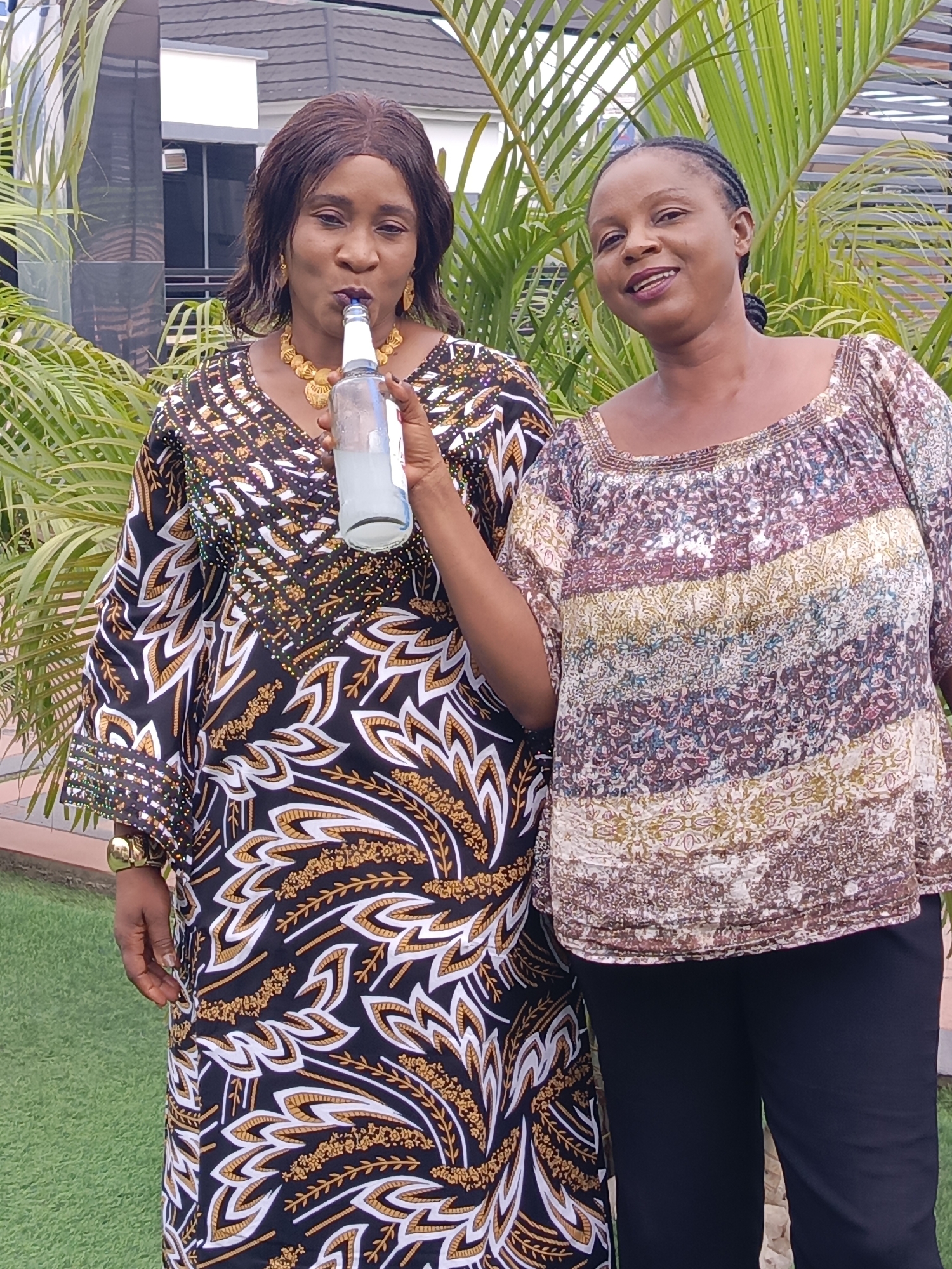 Two women are captured outdoors in Akwanga, Nigeria, standing in front of lush green palm foliage and a partially visible modern building. The woman on the left, with dark skin and dark, medium-length hair, wears a long, intricately patterned dress featuring black, white, and gold motifs. She is adorned with a prominent golden necklace and matching earrings. She holds a clear glass bottle containing a light-colored liquid, with a blue cap, to her mouth and appears to be drinking from it. Her gaze is directed towards the bottle.
To her right, the second woman, also with dark skin and dark braided hair pulled back, looks towards the camera with a subtle smile. She is dressed in a loose-fitting, multi-colored top with patterns in shades of brown, white, blue, green, and gold, paired with dark trousers. The ground beneath them is covered with artificial green grass. The scene is brightly lit, indicating it is daytime. Both women appear relaxed. No visible text is present in the image.