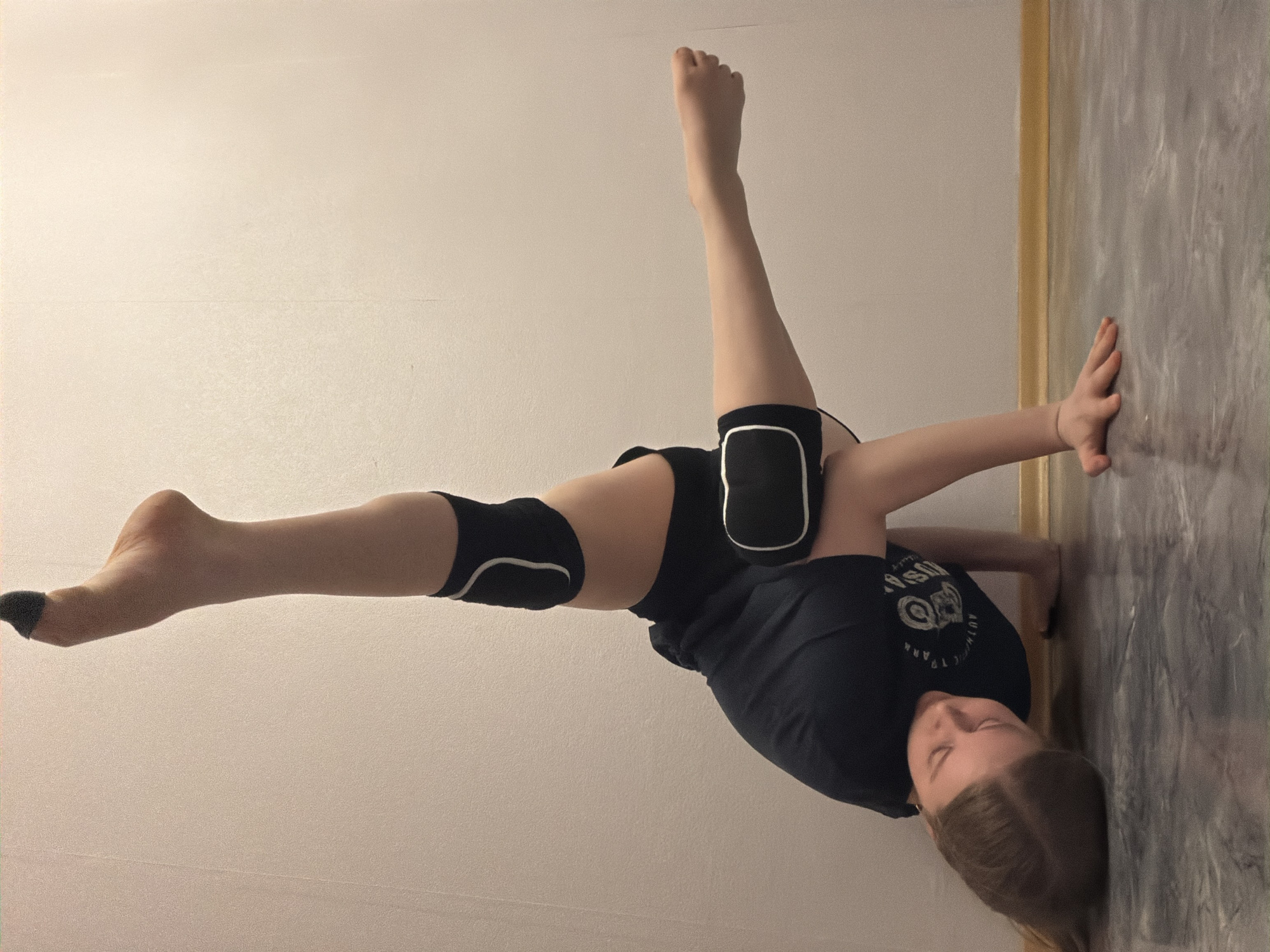 A person is performing a gymnastic or acrobatic maneuver in an interior space. The person is leaning against a wall with one hand while balancing on their stomach and one arm. The legs are spread in different directions, one extending straight out and the other reaching vertically upwards. The person is wearing black shorts, a dark t-shirt, and knee pads. The background shows a plain wall on one side and a patterned wall on the other. There is no information to suggest this scene is in Perm, Russia.