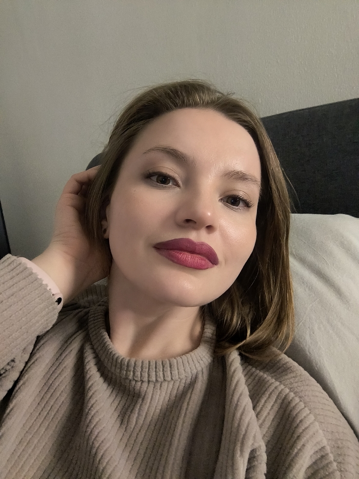 A woman poses close to the camera, her hand near her ear, resting on a bed. She has fair skin, light brown hair, and wears a ribbed beige sweater. The focus is on her face, highlighting her dark eyes and bold raspberry lipstick. She appears relaxed and comfortable, likely at home. The background is muted, with a dark headboard and a light-colored pillow partially visible.