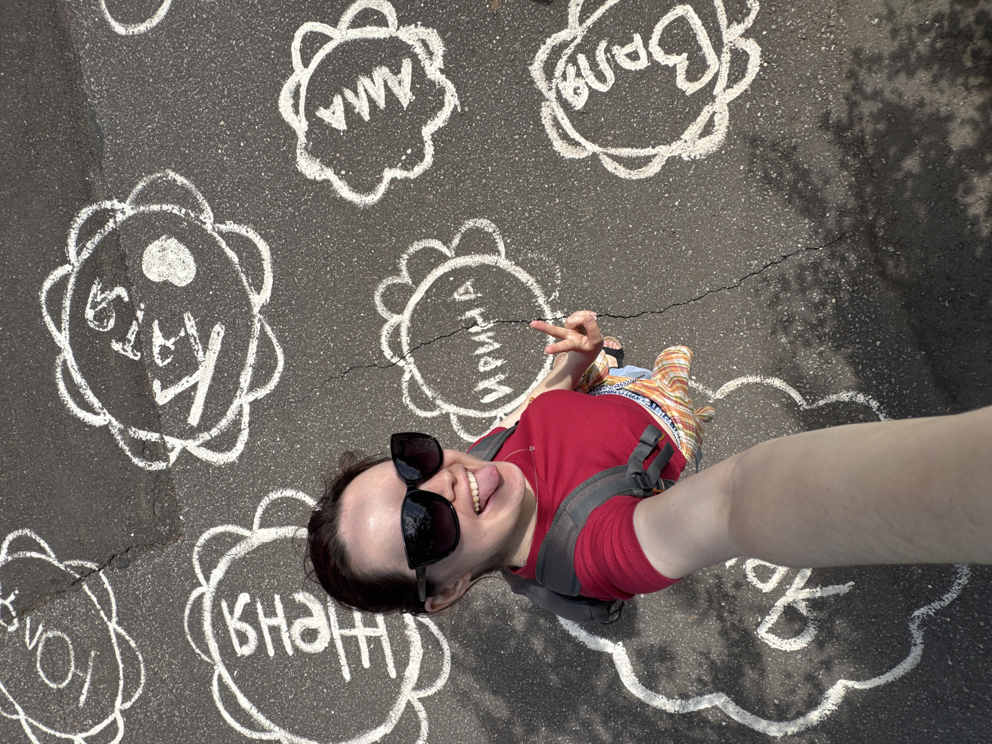 The image displays a high-angle selfie taken on asphalt pavement, identified as being in Izhevsk, Russia. The pavement surface is dark gray, marked by several cracks, and covered with white chalk drawings.
In the foreground, a single adult female is visible from the chest up, taking the photo with an extended left arm. She wears dark-rimmed sunglasses, a red short-sleeved top, and the straps of a gray backpack are visible over her shoulders. A delicate heart-shaped pendant hangs from a thin chain around her neck. Her mouth is open with her tongue extended. Her right hand, positioned in the lower center-right, forms a V-sign with the index and middle fingers extended. She wears a plaid garment, possibly shorts or a skirt, visible below her red top.
The asphalt pavement features multiple white chalk drawings, each enclosed within a multi-lobed, flower-like or cloud-like border. Each drawing contains a Cyrillic name. Visible names include:
* "ЮЛЯ" (YULYA) in the upper left.
* "ЖЕНЯ" (ZHENYA) in the middle left.
* "КАТЯ" (KATYA) with a central heart symbol in the upper center.
* "ИРИНА" (IRINA) in the middle right.
* "АНЯ" (ANYA) partially visible in the upper far right.
* "ВЛАДА" (VLADA) with a central heart symbol in the lower right.
* "ЯНА" (YANA) partially visible in the lower left.
Bright sunlight illuminates the scene, casting distinct shadows on the pavement, including a mottled shadow of tree foliage in the lower right quadrant.