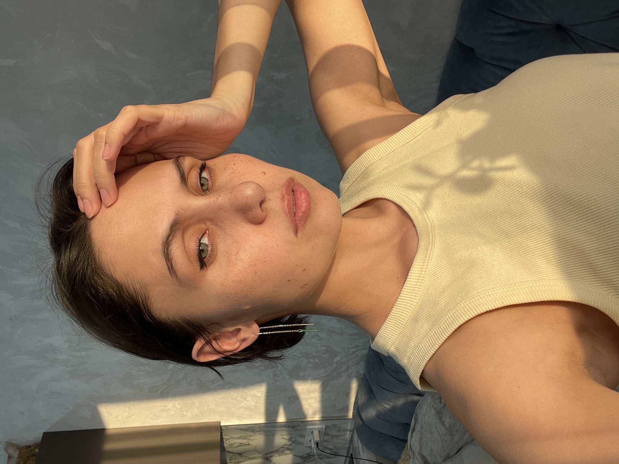 A close-up, correctly oriented portrait features a young woman reclining in an indoor setting, looking directly at the camera with a calm expression. Her right hand is gently placed on her forehead.
She has fair skin with visible freckles across her face and possesses striking green-hazel eyes. Her dark hair is styled short or pulled back, revealing a distinctive ear cuff or earring on her left ear, featuring multiple delicate chains or bars. She is wearing a light yellow or cream-colored ribbed tank top. Her lips are slightly parted, showcasing their natural pink color.
Strong, directional sunlight illuminates the scene, casting prominent linear shadows across her arms and chest. Geometric shadows are also visible on the textured grey wall behind her and on a light-colored surface below, which features a marble-like pattern. This lighting suggests either early morning or late afternoon.
The background details include a dark blue upholstered surface (likely a sofa or bed) behind her upper body, and a white electrical outlet with a plug on the lower right, next to a dark rectangular object. The setting appears to be an interior space, possibly near a window, in Izhevsk, Russia. There is no visible text in the image.