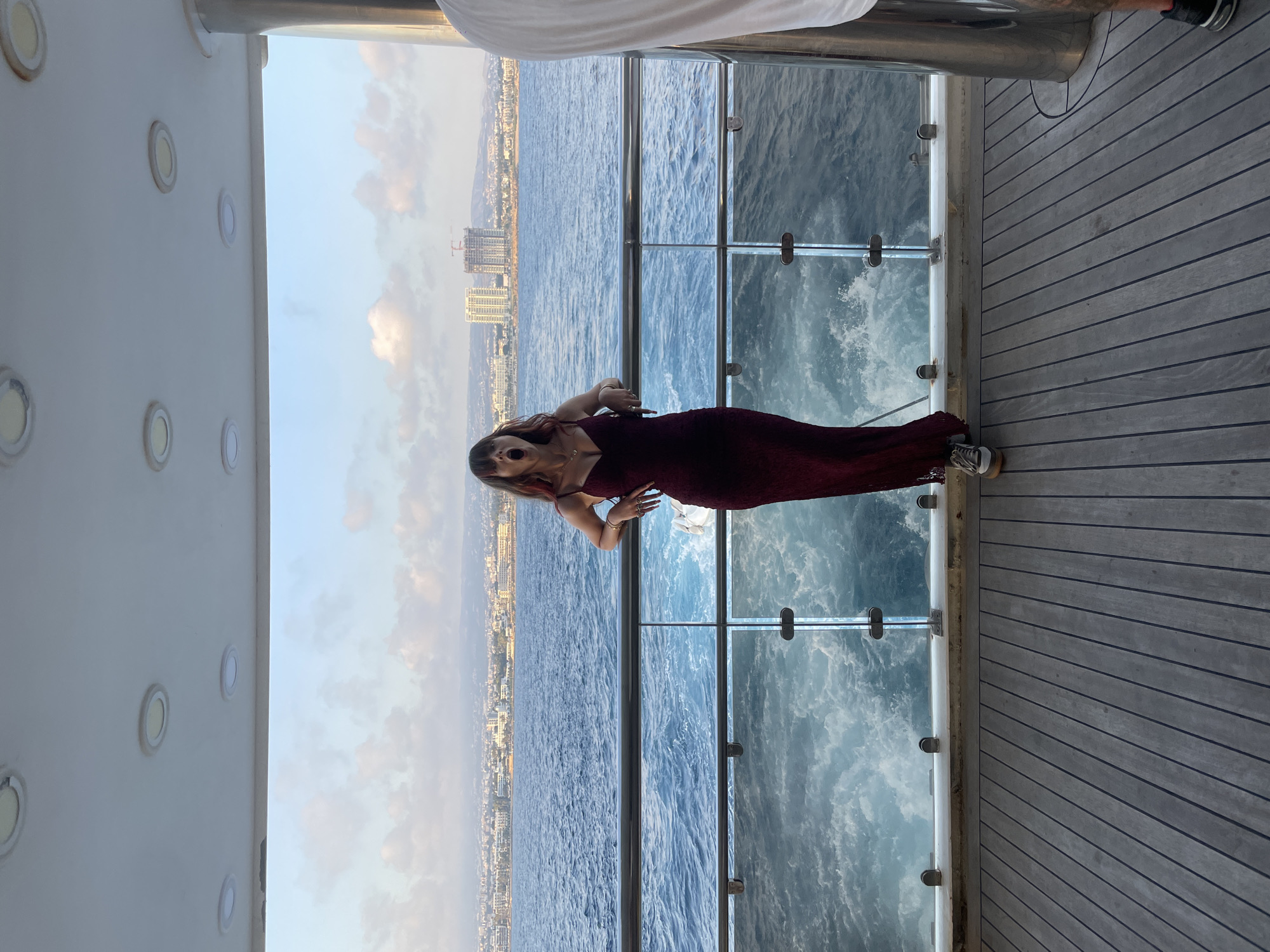 A young woman with reddish-brown hair, wearing a long, burgundy, lace dress and grey sneakers, stands on a wooden deck. She is leaning on a glass railing overlooking the sea, with her mouth wide open in a yawn. Behind her, a coastal city with buildings and construction cranes is visible under a cloudy sky. The sea is a deep blue with white waves churning. The setting appears to be a luxury yacht, possibly docked or moving in the waters near Paphos Municipality, Cyprus. Another person, wearing a white shirt, is partially visible on the right side of the frame.