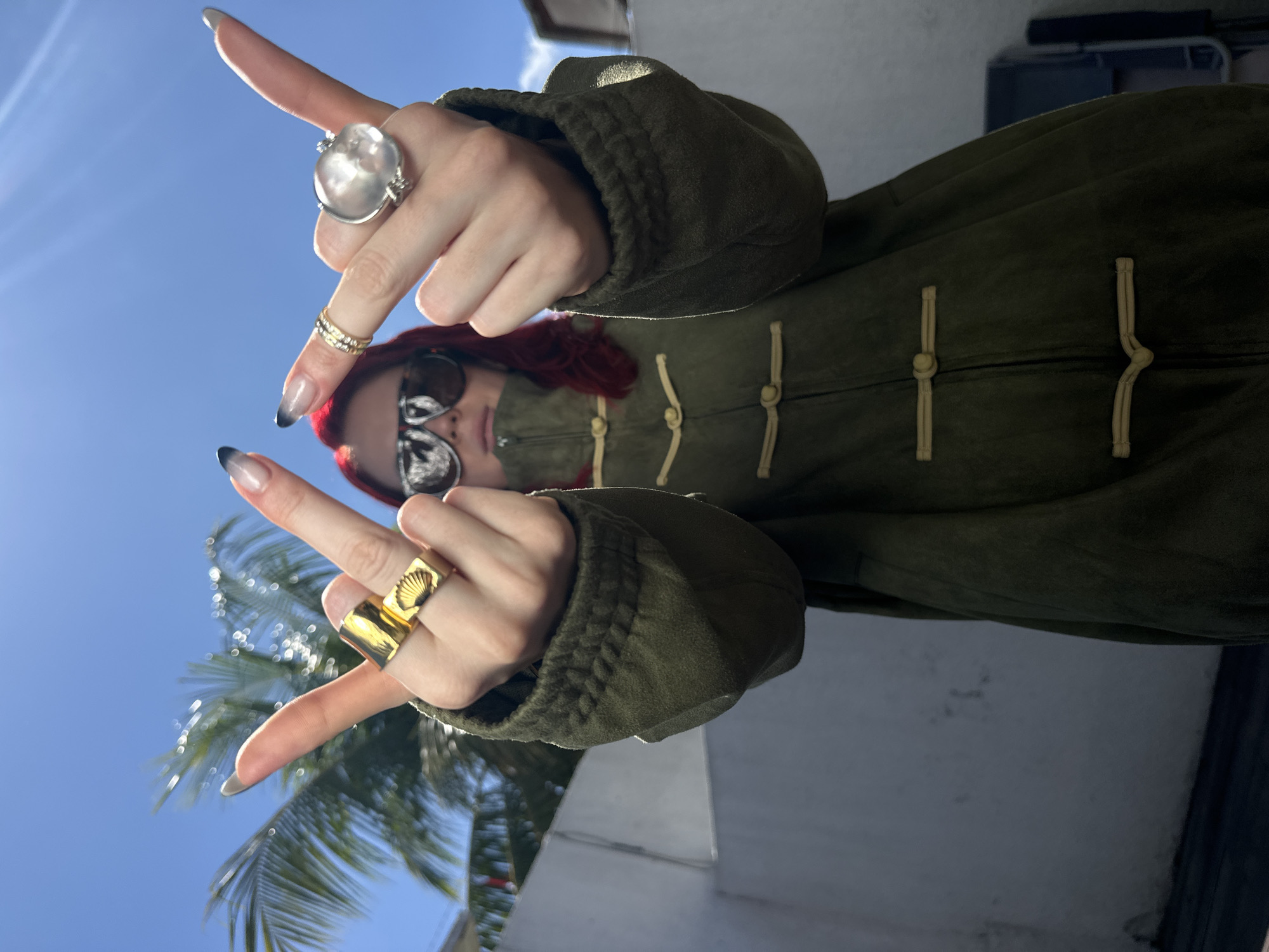 A low-angle, outdoor, medium shot captures a person with vibrant red hair, wearing dark sunglasses and a green, Chinese-style coat with toggle closures, against a bright blue sky and a palm tree. The individual is looking up at the camera, holding both hands up with middle fingers extended. On the person's left hand, two prominent gold rings are visible: a wide, flat band with a carved shell design and a thinner, textured band. On the right hand, a delicate gold band sits above a large, clear, irregular-shaped stone set in a silver ring. The background shows a white wall and some foliage, suggesting a tropical or subtropical urban environment, possibly within the city of North Kuta, Indonesia. The lighting is strong, casting shadows and creating a bright, sunny atmosphere.