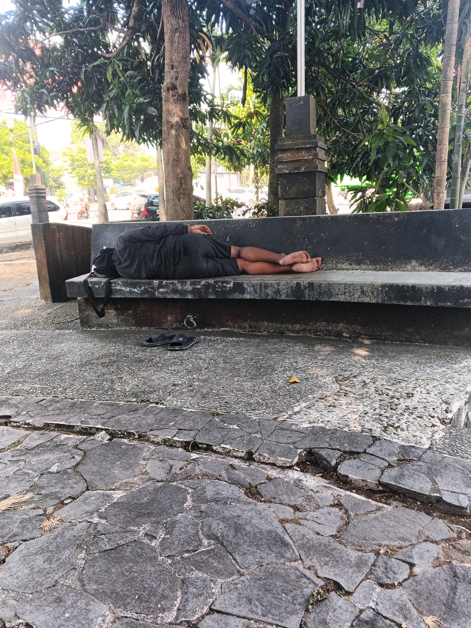 A person is sleeping on a stone bench outdoors, possibly in a public park or plaza in Ungaran, Indonesia. The individual is lying on their side, facing away from the camera, and is covered in dark clothing. Their feet are visible, and a pair of black flip-flops are placed on the ground next to the bench. The bench itself is made of dark, weathered stone, and is situated on a patterned stone ground. Trees with green foliage are visible in the background, along with some traffic and buildings, suggesting an urban or semi-urban environment. The lighting indicates it is daytime.