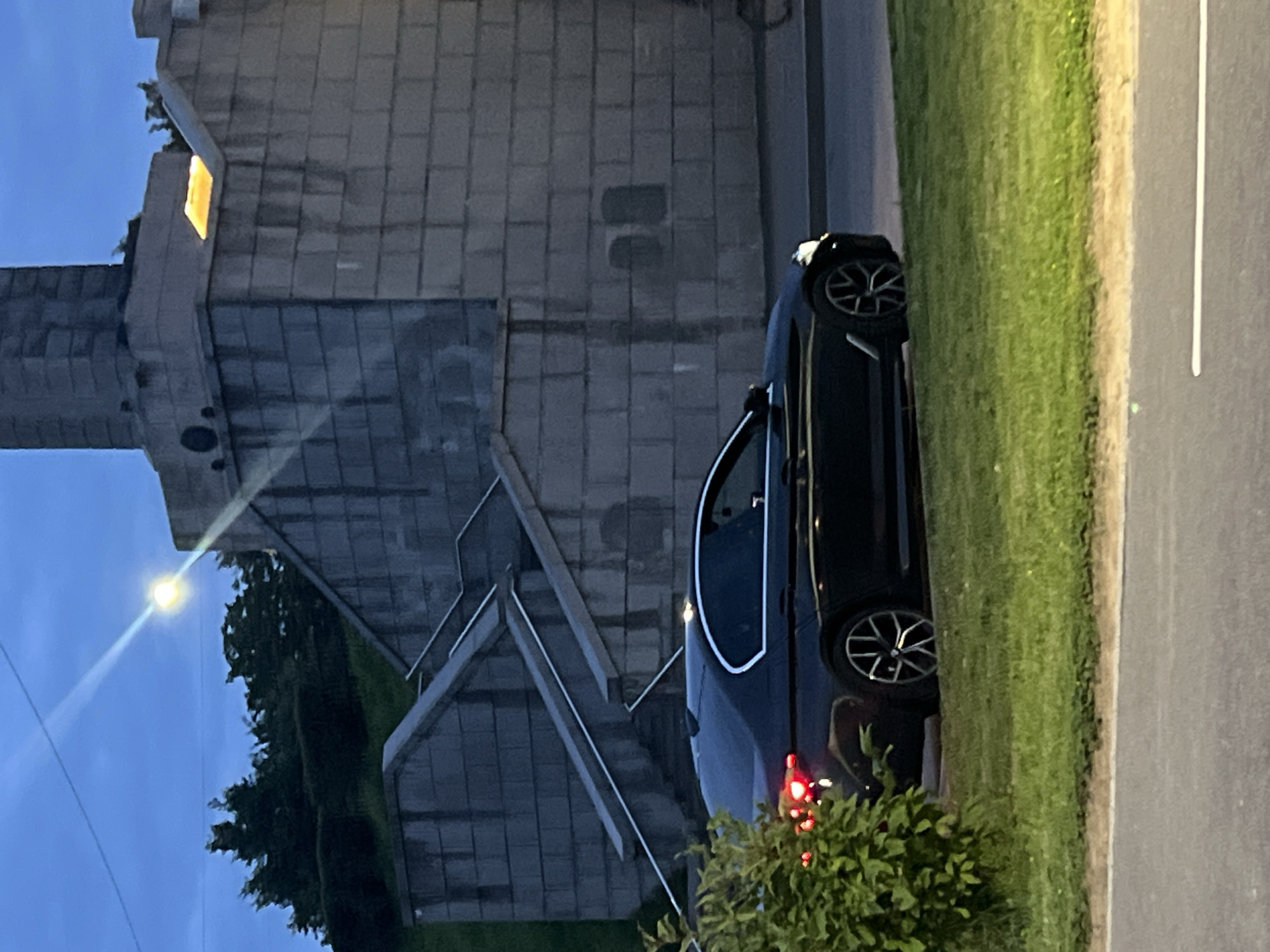 A black sedan is parked on a grassy area next to a paved road, with its rear brake lights illuminated. In the background, a monumental stone structure with visible staircases and a light source on its upper level dominates the scene. Trees are visible on a grassy incline to the left of the structure. The sky is dark blue, indicating either dusk or dawn, with a bright light source visible in the upper left quadrant of the image, possibly a streetlamp or a powerful distant light. The overall setting suggests an outdoor urban environment.