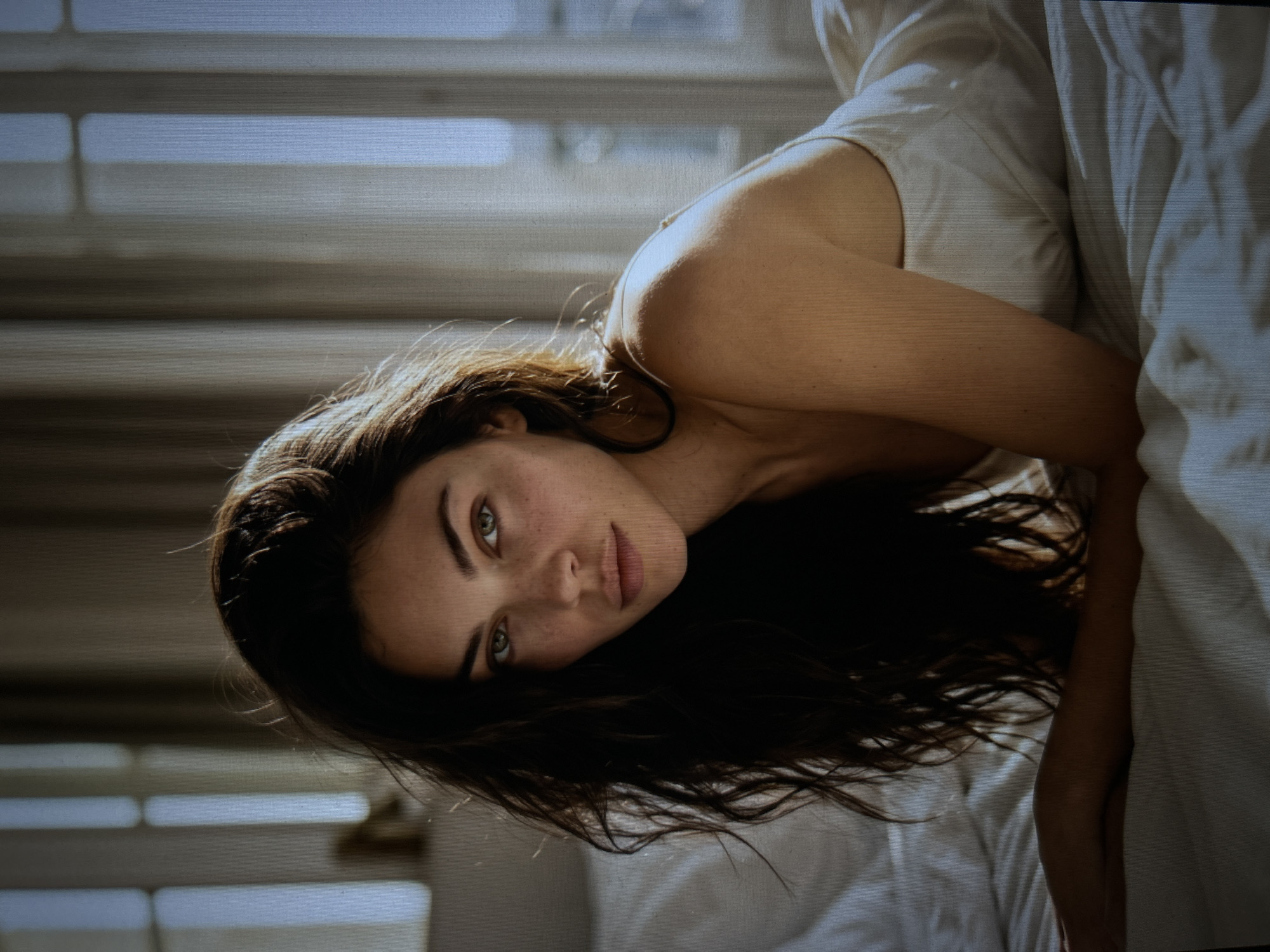 A woman with dark, wavy hair and freckled skin is lying on a bed. She is facing the camera, with her head tilted slightly to the left. Her eyes are green, and her lips are full and slightly parted. She is wearing a white slip. The background is blurred, but appears to be a room with large windows and sheer curtains. The lighting is soft and natural, coming from the windows. The overall impression is one of intimacy and vulnerability.