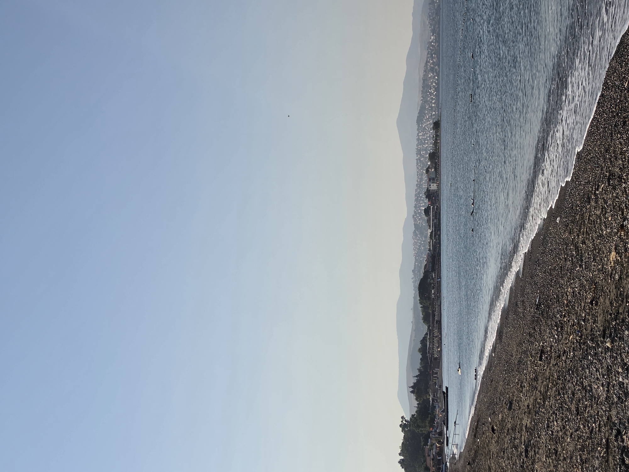 This is a wide shot of a rocky beach and the Aegean Sea, taken from a low angle. The foreground is dominated by the dark, pebble-strewn beach, with a gentle wave washing ashore, creating a thin line of white foam. Beyond the beach, the calm blue sea stretches to the horizon, dotted with a few small buoys or markers forming a line parallel to the shore. In the middle ground, a town or village is visible along the coastline, with buildings nestled amongst trees on a gentle slope. Further in the distance, rolling hills and mountains form a hazy backdrop against the clear, pale blue sky. There are no people clearly visible in the image, though there might be a few distant figures on the beach or in the water. The overall impression is one of a peaceful coastal scene. The location is likely Seferihisar, Turkey, a coastal town known for its scenic beauty and proximity to the Aegean Sea.