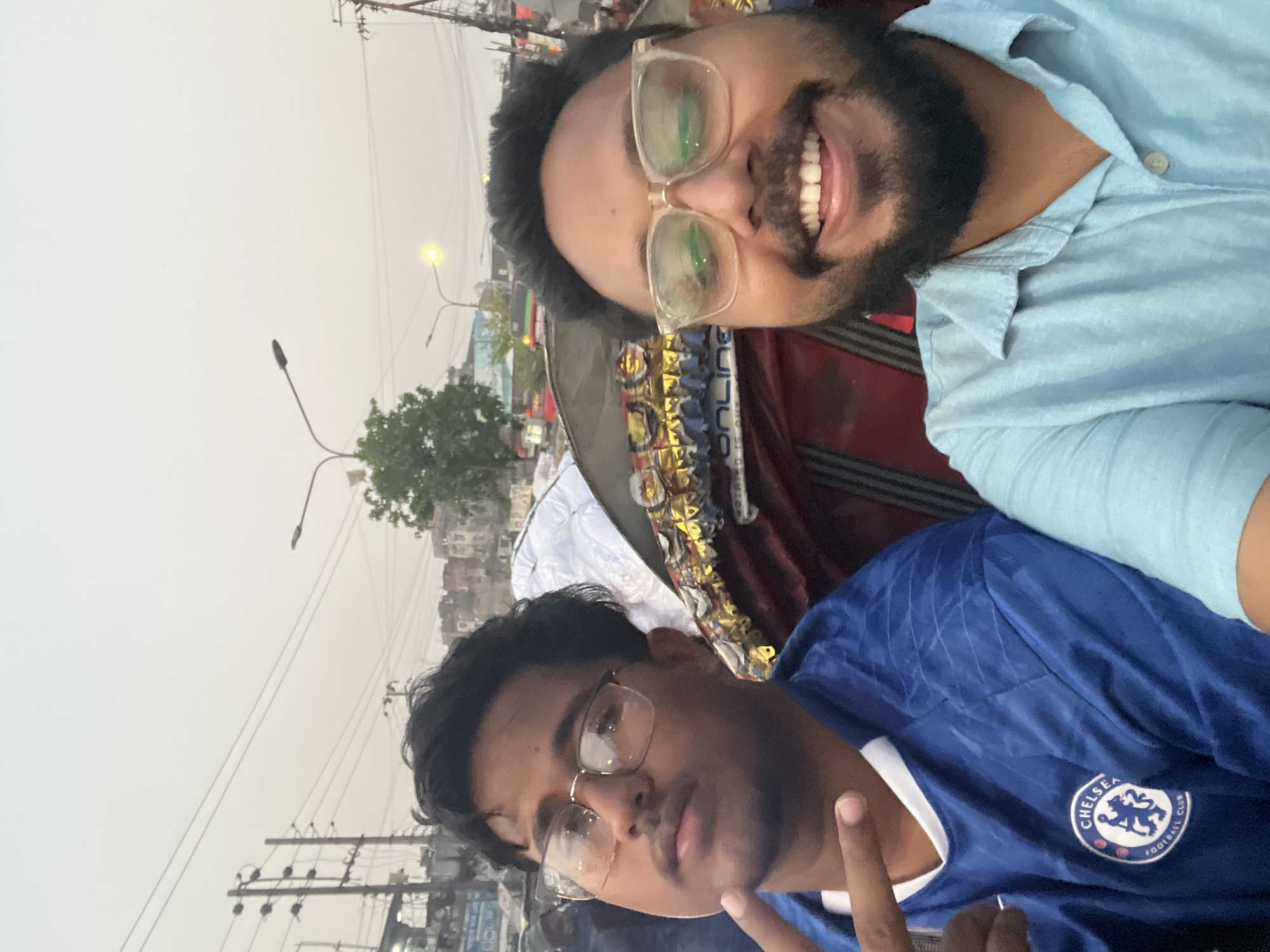 Two men are in a rickshaw, likely traveling on a street in Dhaka, Bangladesh. One man is wearing a blue Chelsea football jersey, and the other a light blue button-down shirt and glasses. Buildings, power lines, and street lights are visible in the background. The sky appears overcast.