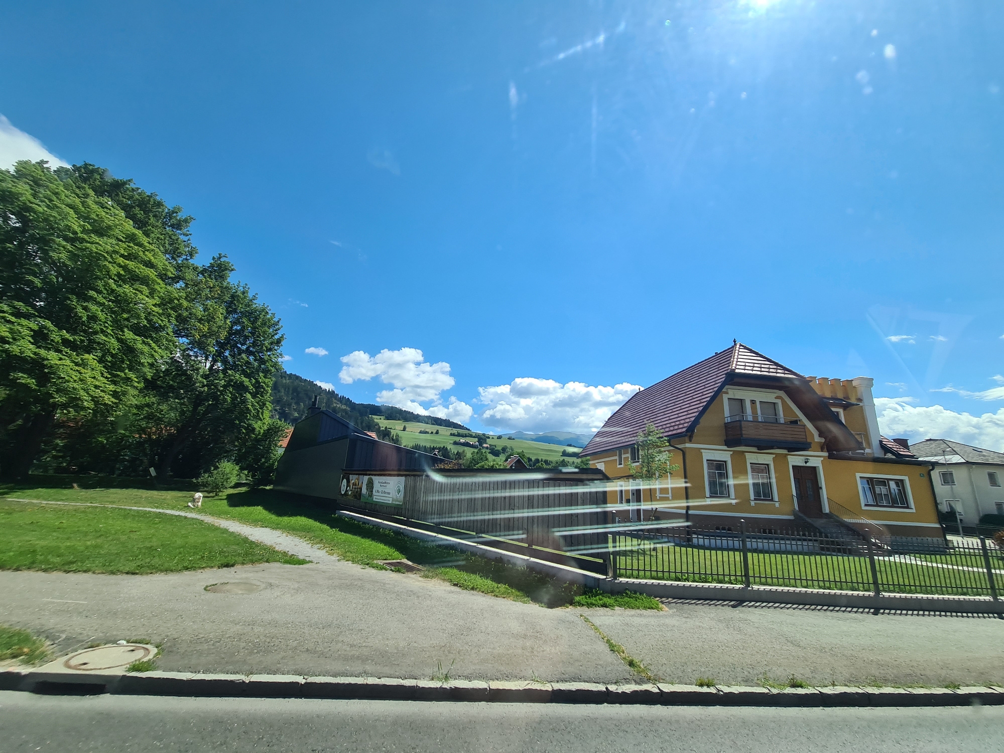 The image shows a residential area on a sunny day with blue skies and scattered clouds. In the foreground, there is a paved road with a curb and a manhole cover. To the left of the road is a grassy area with a path winding through it. Several trees with lush green foliage are on the left side of the image, and a small, light-colored animal, possibly a dog, is visible on the grass near the trees.
In the middle ground, there are several buildings. A long, low building with a dark roof and wooden siding is prominent. Adjacent to it is a larger, two-story house painted yellow with a reddish-brown tiled roof. This house has multiple windows, a balcony on the upper floor, and a dark wooden door with a set of stairs leading up to it. A black metal fence runs along the front of the yellow house and a portion of the darker building.
In the background, rolling green hills with some scattered houses and trees can be seen, suggesting a semi-rural or suburban setting within the city of Sankt Marein bei Neumarkt, Austria. The sun is shining brightly, casting a slight glare on the image, particularly in the upper right corner. The overall scene conveys a peaceful, pleasant day in a scenic Austrian location.
