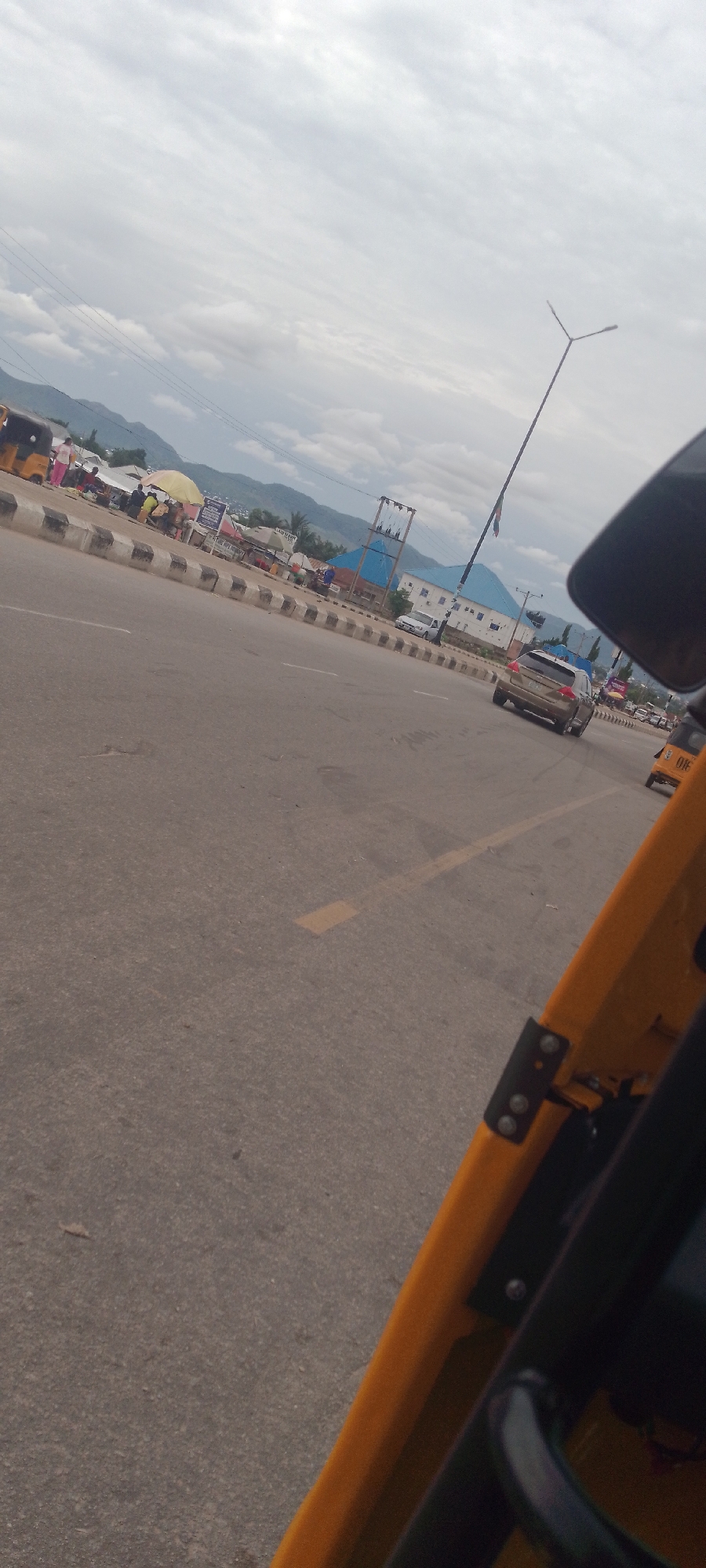 The image depicts a street scene in Jalingo, Nigeria, likely in a market area adjacent to a road. In the foreground, a portion of a yellow vehicle, possibly a tricycle or auto-rickshaw, is visible. The road itself appears to be asphalt with painted lane markers. Along the side of the road, there is a raised sidewalk or median with concrete and black and white striped barriers.
Beyond the barriers, a bustling market area is apparent with various stalls and structures, some covered by umbrellas. Several people are present, engaged in what appears to be market activities. A yellow tricycle is parked on the left side. In the mid-ground, vehicles are on the road, including a light-colored sedan and a darker SUV. In the background, buildings with blue and white roofs are visible, and further beyond, rolling hills and mountains are silhouetted against a cloudy sky. A tall, slender street light is prominent in the upper portion of the image. The overall atmosphere suggests a busy urban environment.