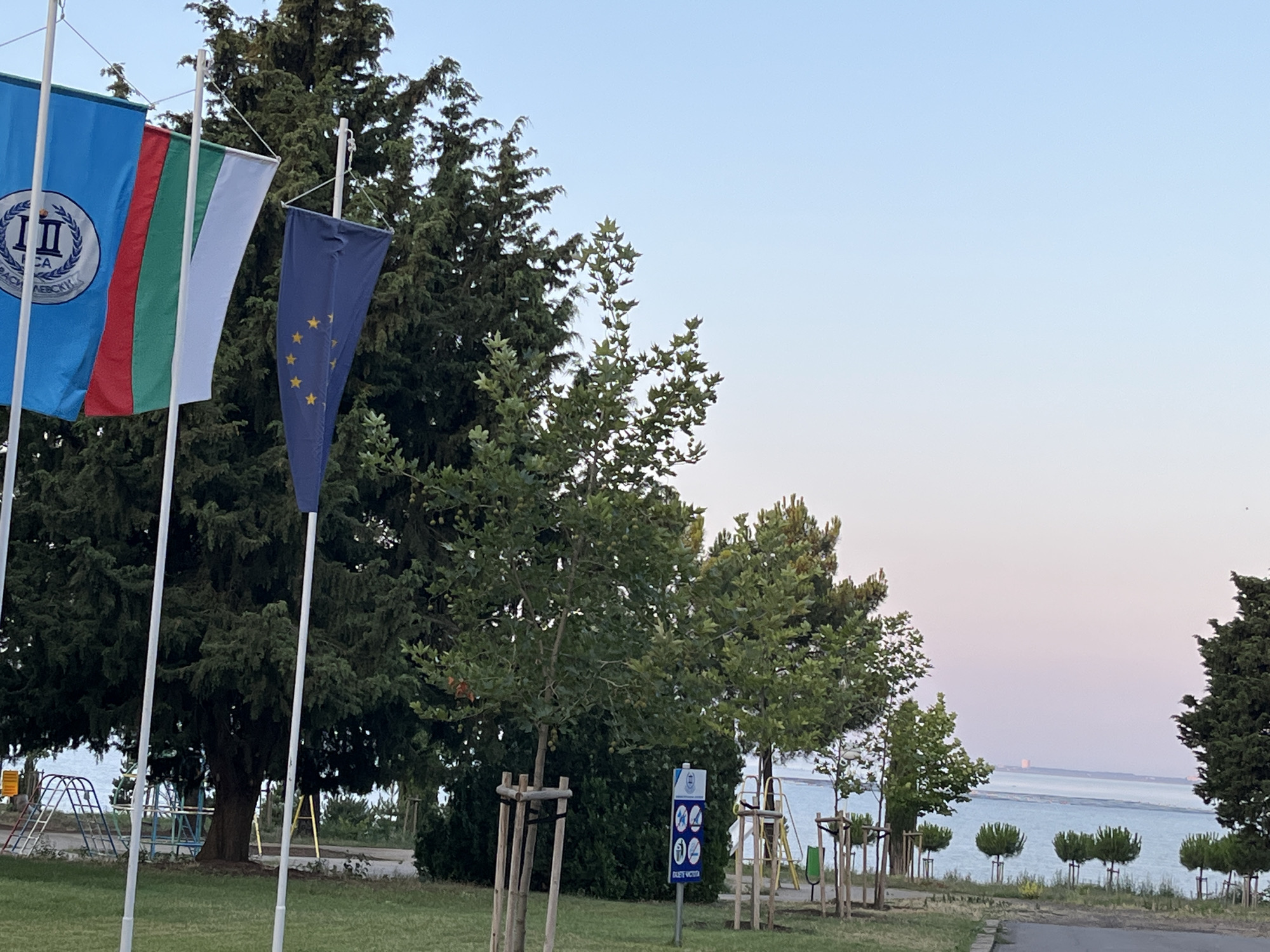 The image depicts a waterfront scene in Nesebar, Bulgaria, under a clear, pale blue sky that transitions to a lighter hue near the horizon. In the foreground, several flags are prominently displayed on white poles. The leftmost flag is blue with a circular emblem containing text. Next to it are the Bulgarian national flag, featuring horizontal stripes of white, green, and red. A dark blue flag with yellow stars, likely representing the European Union, is also visible.
Behind the flags, a dense evergreen tree dominates the left side of the frame, with lighter green deciduous trees situated to its right. The ground is covered in grass, with a paved path running diagonally from the lower right corner.
Further into the scene, several young trees with visible wooden supports are planted. A sign with multiple icons and text is present near these trees. In the background, a body of water, presumably the sea, stretches to the horizon. Several small, rounded trees are planted along the water's edge. In the distance, across the water, a hazy structure is visible. Playground equipment, including swings and a climbing frame, can be seen on the left side of the image, nearer the water. The overall atmosphere is tranquil, with no people present.