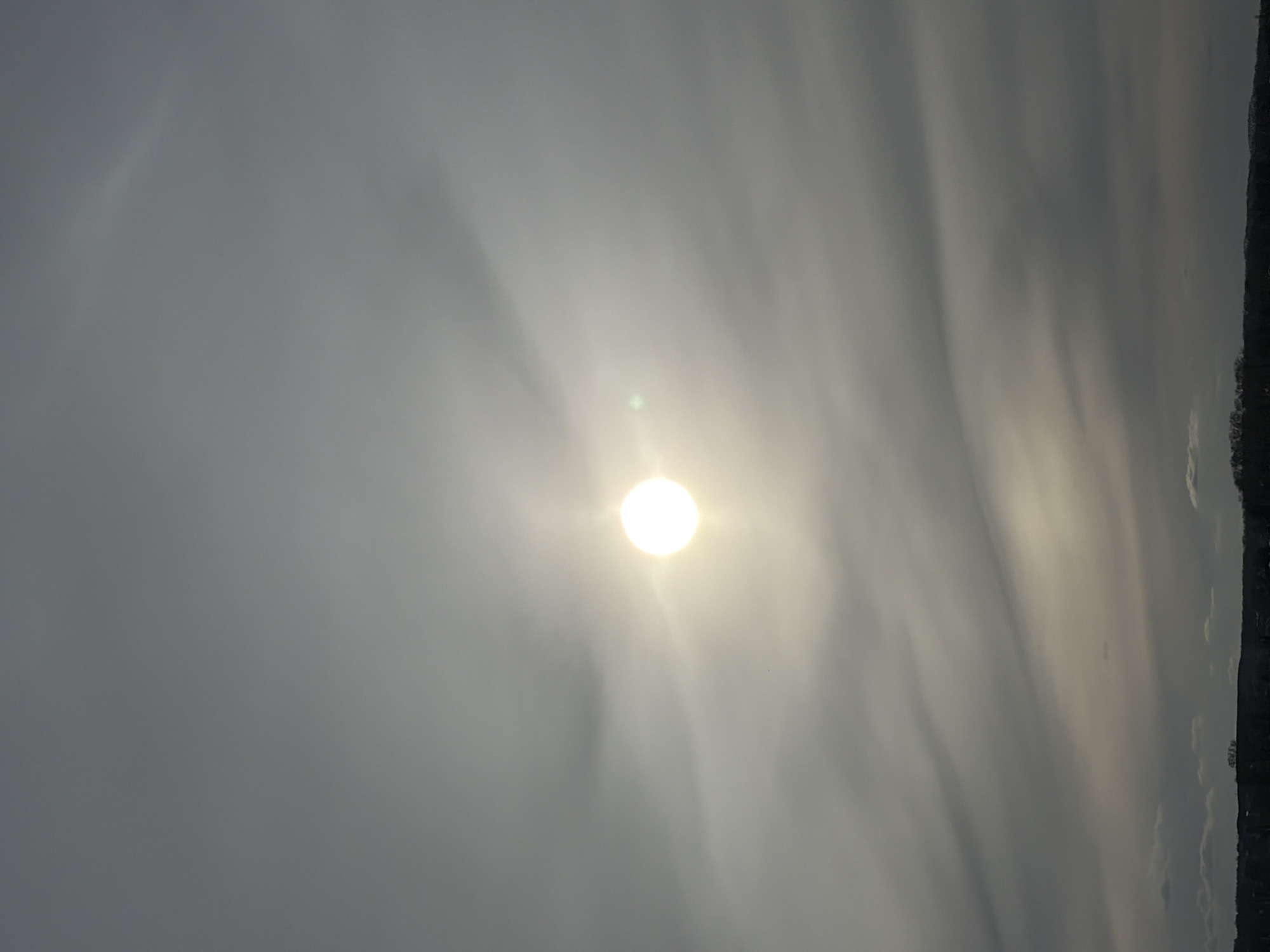 The image depicts a hazy sky with the sun visible through the cloud cover. The brightness of the sun is concentrated in the center. The sky is grey and cloudy. There is an indistinct treeline on the right edge of the frame. The image is rotated. Without additional information, it's impossible to confirm if the image was taken in Votkinsk, Russia.
