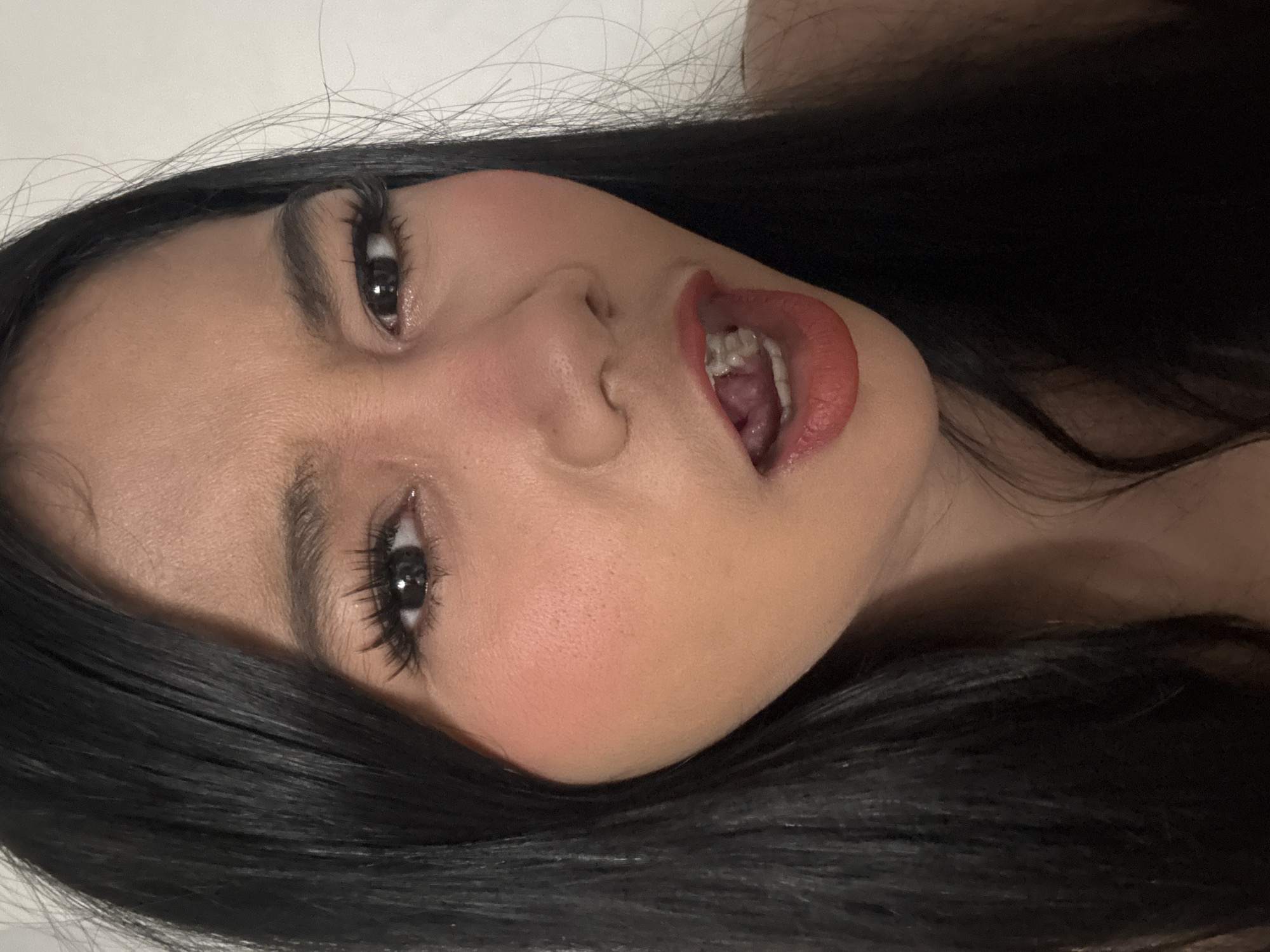 A close-up, headshot of a young woman is depicted. She has long, dark hair framing her face, which is turned slightly to the side. Her eyes are wide open, with prominent eyelashes, and her eyebrows are neatly shaped. Her nose is slightly tilted, and her lips are parted, revealing her teeth. She is wearing coral-colored lipstick and blush. The background is a plain, light-colored wall. This image was taken in North Jakarta, Indonesia.