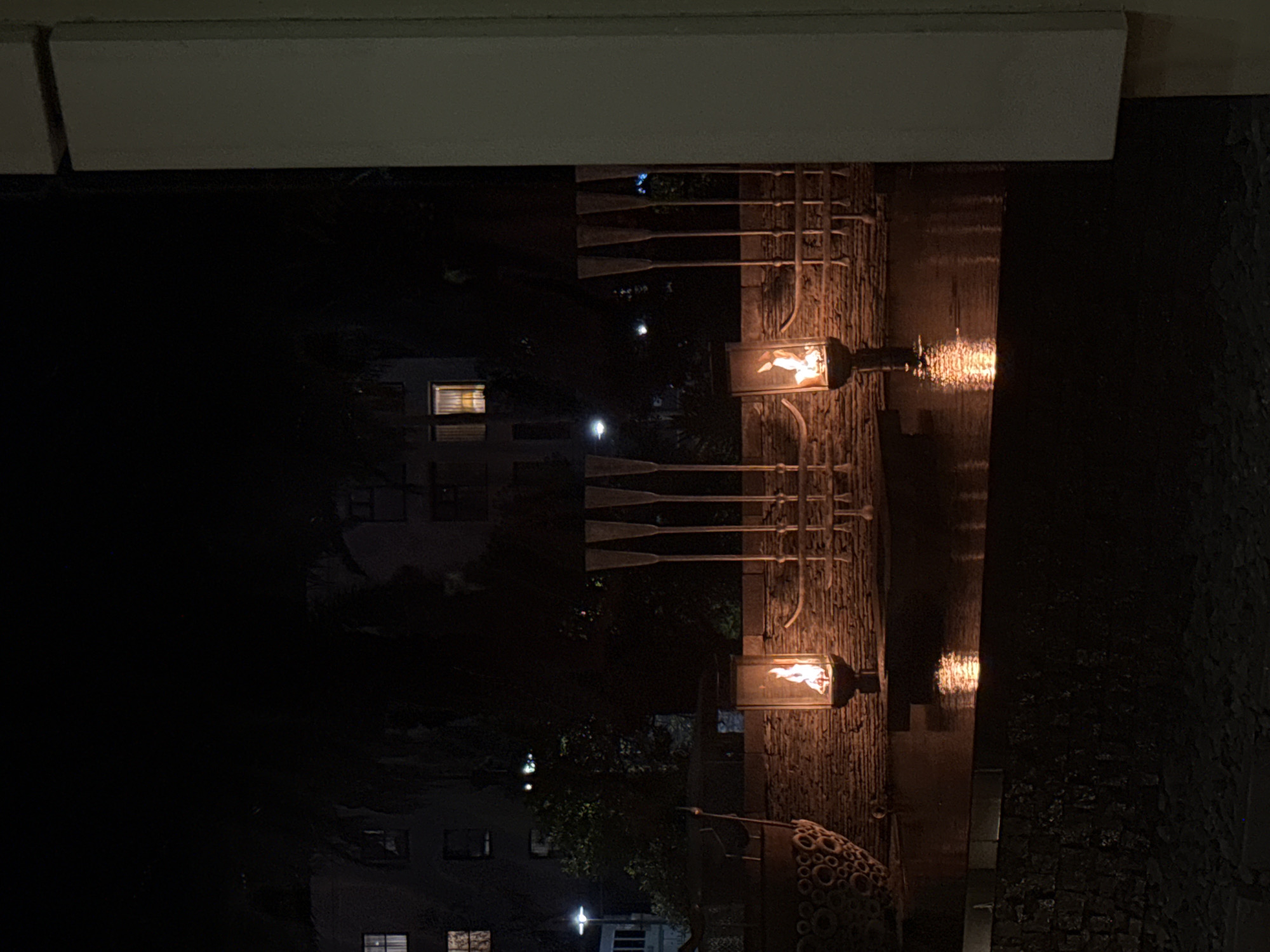 The image shows a nighttime scene in Cape Town, South Africa. A low-angle perspective captures a building exterior, likely a hotel or upscale establishment, featuring decorative metalwork resembling oars extending from a stone wall. Two gas lamps, containing visible flames, illuminate the wall and a flowing water feature that creates a reflective surface. In the background, the silhouette of a multistory building with illuminated windows suggests a residential or commercial area. The lighting is dim, highlighting the contrast between the flames and the surrounding darkness. A portion of the ceiling or overhang is visible in the upper part of the image. The overall atmosphere is upscale and quiet.