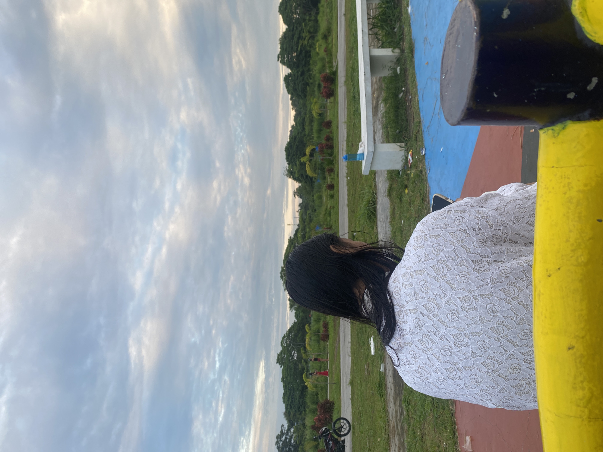The image, oriented horizontally, depicts a person from behind, positioned on the right side of the frame, looking out towards an outdoor park scene. The individual has long, dark hair and is wearing a white, intricately patterned lace top.
In the foreground on the far right, a yellow pole or structure is prominent, with a dark, cylindrical top. Adjacent to this, the ground is painted with sections of reddish-brown and blue, indicating a recreational area. Further into the midground, a paved pathway runs through grassy areas, lined with scattered trees and shrubs. A white concrete bench with a blue water bottle placed upon it is visible next to the path. In the lower left, part of a bicycle or motorcycle wheel can be seen.
The background features a dense line of green trees against an expansive sky filled with soft, light blue and white clouds, suggesting either an early morning or late afternoon setting with gentle, diffused light. Some litter is visible on the grass and near the foreground structures. There is no visible text in the image.