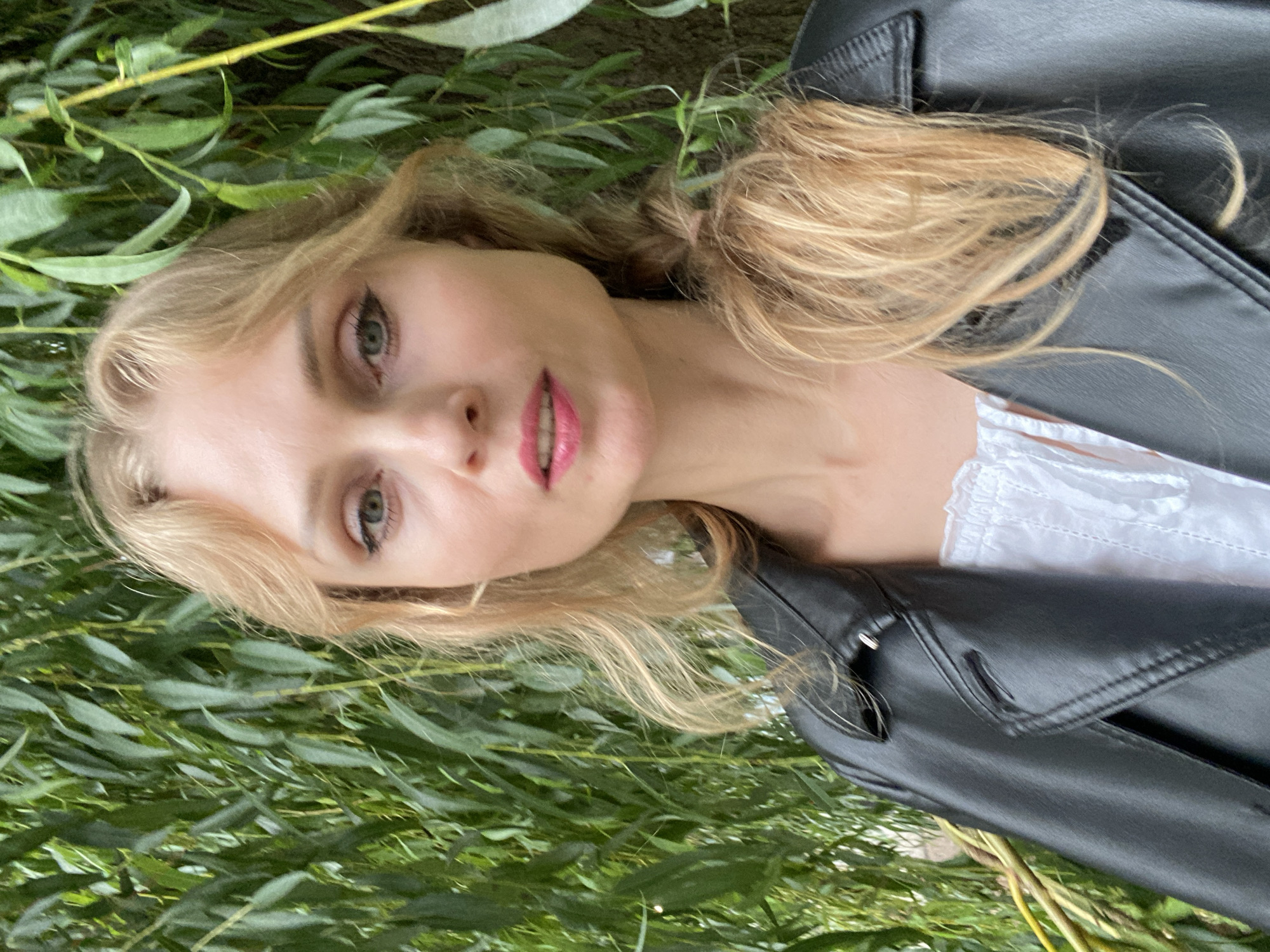 A portrait of a blonde woman wearing a black leather jacket and a white top is shown outdoors. She has light green eyes, fair skin, and her hair is a medium blonde color, styled loosely around her face and pulled back into a ponytail. She is looking directly at the camera with a neutral expression. The background is a dense foliage of green leaves, suggesting a park or garden setting. The lighting is soft and natural, indicating it was taken during the daytime. The image does not contain any information or visual cues that would place it specifically in Lida, Belarus.