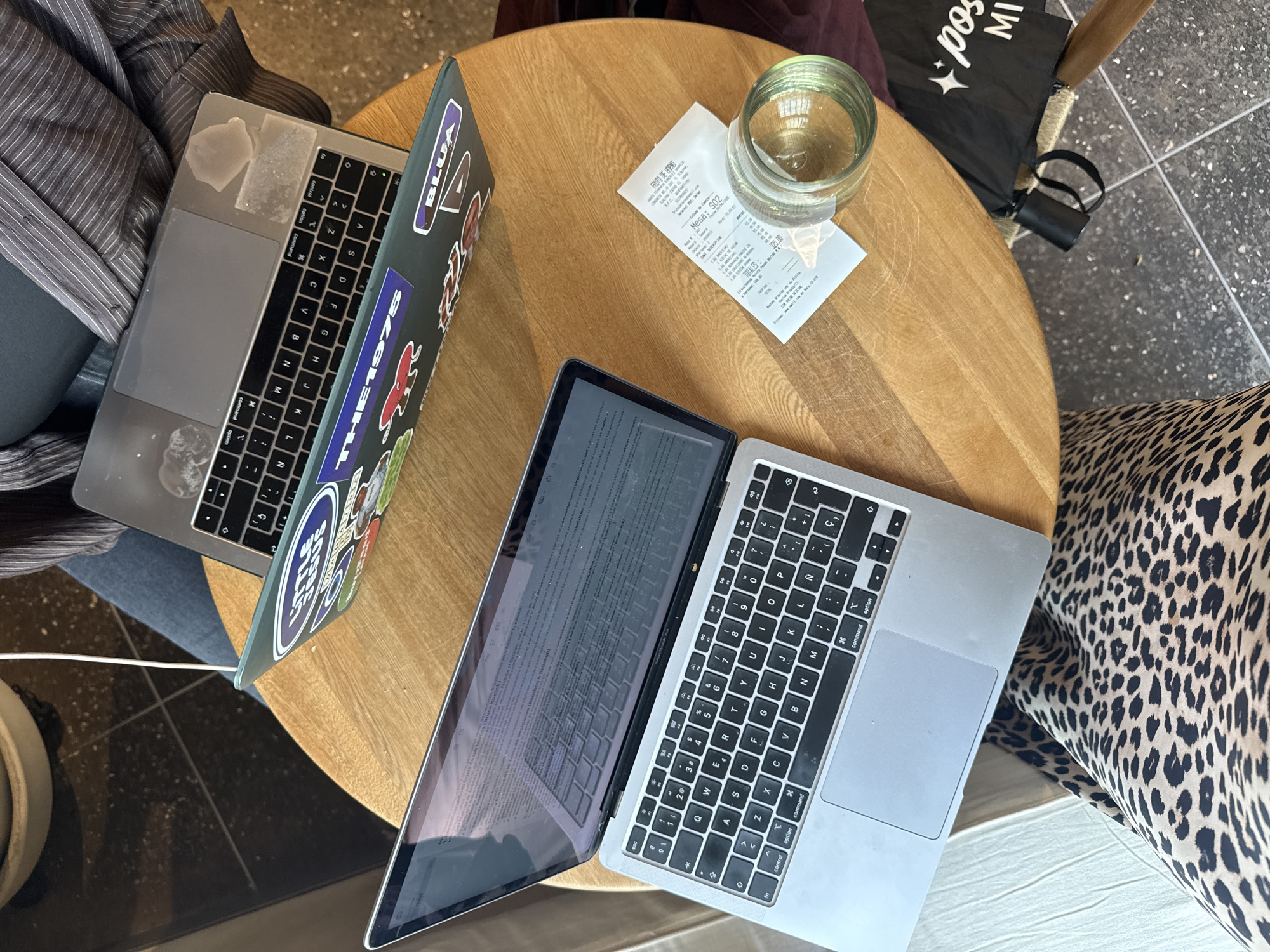 The scene is a close-up, overhead shot of a circular wooden table in what appears to be a cafe or workspace in Mexico City, Mexico. Two open laptops are prominently featured, one with stickers on its lid. A glass of water and a receipt sit on the table next to the laptops. Part of a chair with a black-and-white patterned cushion is visible on the right. On the left, a person is seated with a striped garment. A black tote bag leans against the chair in the background.