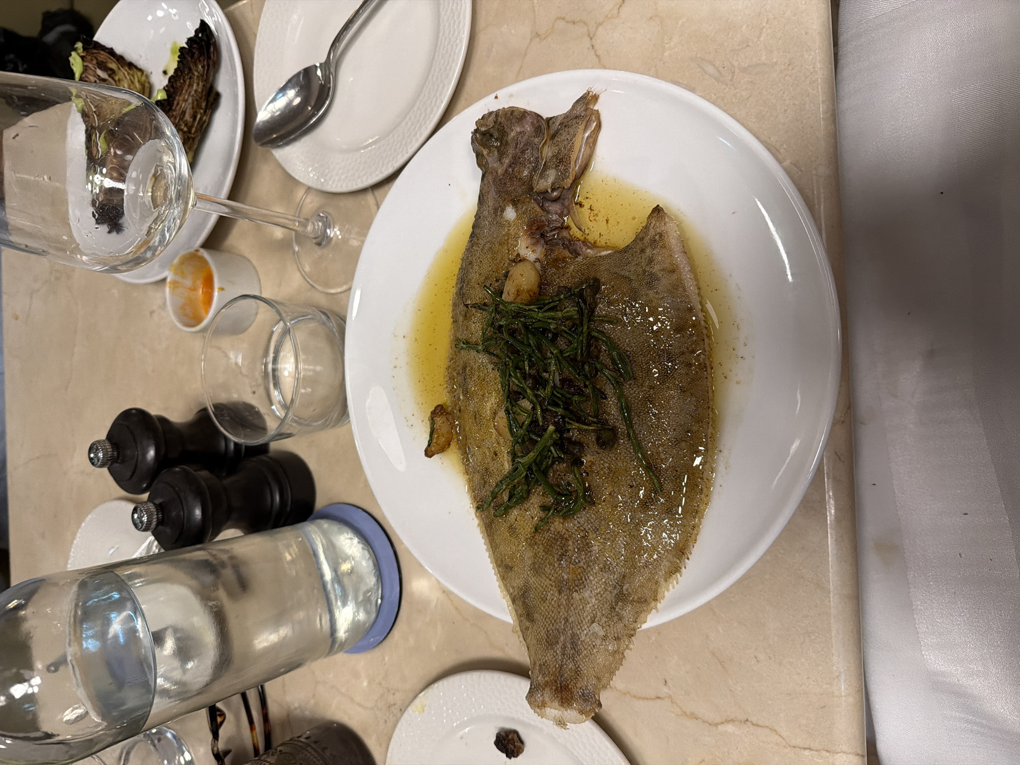 The image shows a table setting at a restaurant, likely in London, United Kingdom, with a prepared meal of pan-seared fish served on a white plate, accompanied by a glass of water, a wine glass, salt and pepper shakers, and a small dish of sauce. The fish, possibly a Dover sole, is golden brown with crispy skin and garnished with green herbs and a slice of garlic. It is swimming in a pool of melted butter or oil. In the background, a partially eaten salad of roasted cabbage wedges is visible on another plate. The table surface is a light-colored marble with a subtle pattern. The overall ambiance suggests a fine dining experience.