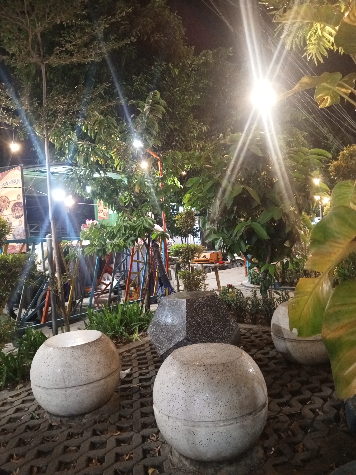 The image depicts an outdoor scene at night in Kloncing, Indonesia, illuminated by bright overhead lights that create strong sunburst effects. In the foreground, three spherical, light-colored stone benches with dark speckles are placed on a patterned gray paving stone surface. Behind these benches, a faceted, dark gray stone object is visible. The middle ground features dense foliage from trees and bushes, with a colorful metal structure resembling a playground or support system behind it. Further back, a tented area with signs and what appears to be food stalls or market stalls is visible, along with more trees and plants. A motorcycle and a small orange vehicle are also present in the background, suggesting a public or commercial space. The overall impression is that of an outdoor gathering area or market operating in the evening.