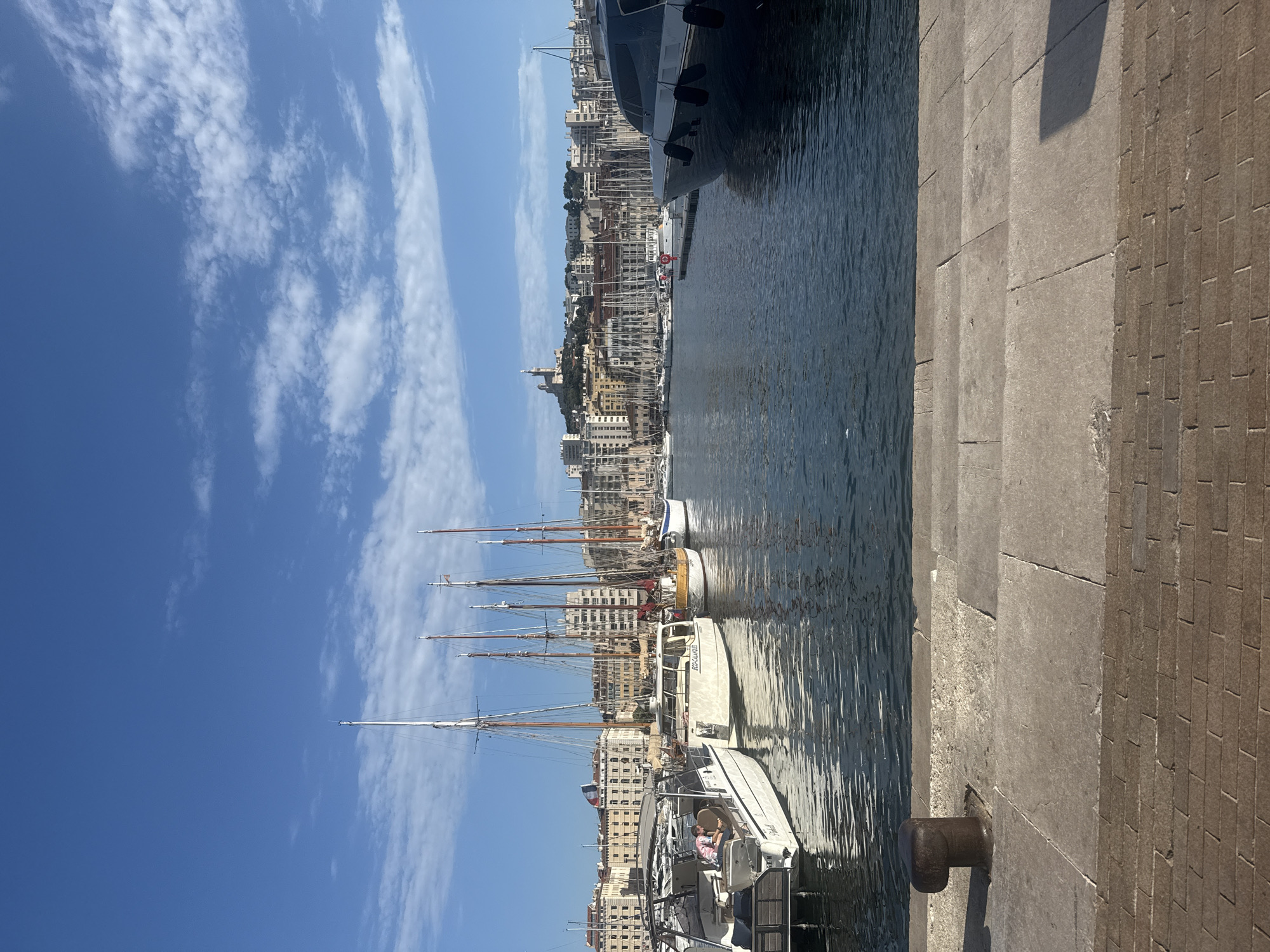 The image depicts a harbor scene under a clear blue sky with scattered white clouds. In the foreground, stone steps lead down to a quay with a metal bollard. The water is calm, reflecting the sky and the boats. A row of boats, including several sailboats with tall masts and a motor yacht, are moored in the harbor. In the background, a city skyline rises, characterized by buildings of varying heights and architectural styles, with a prominent church or cathedral situated on a hill overlooking the city. The overall impression is of a bustling port city on a bright, sunny day.