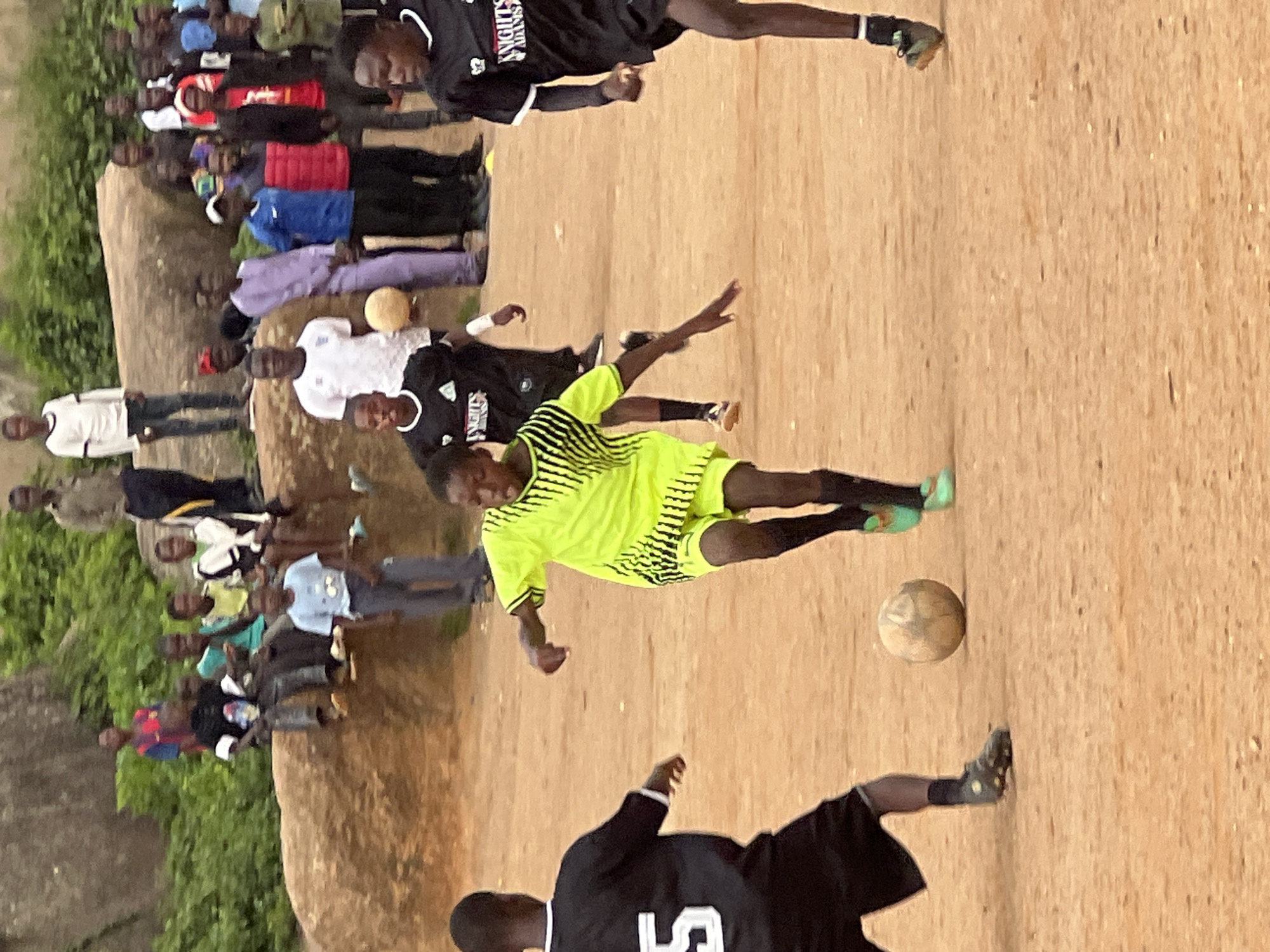 An outdoor, ground-level, medium shot captures a group of young men and boys playing soccer on a dirt field. The foreground is dominated by three young men actively engaged in the game. On the left, a boy wearing a black jersey with the number "5" on the back is positioned with his back to the camera, his right hand outstretched. In the center, a boy in a bright neon yellow jersey with black diagonal stripes and black shorts is in motion, kicking a worn soccer ball forward with his right foot. His left leg is bent, and his left arm is extended backward for balance. To the right, another young man in a black jersey with white trim is running forward, his arms slightly out.
In the middle ground, several more individuals are present, some watching the game and others standing around. A boy in a black and white jersey is visible further back, and another in a black jersey is to his left. In the background, a larger group of spectators, a mix of men and boys, are gathered on and around a large, rough-textured rock formation covered in sparse green vegetation. The overall environment suggests a casual game of soccer taking place in an arid or semi-arid area, consistent with the terrain found in Jos, Nigeria. The lighting indicates daylight, though the sky is not visible.