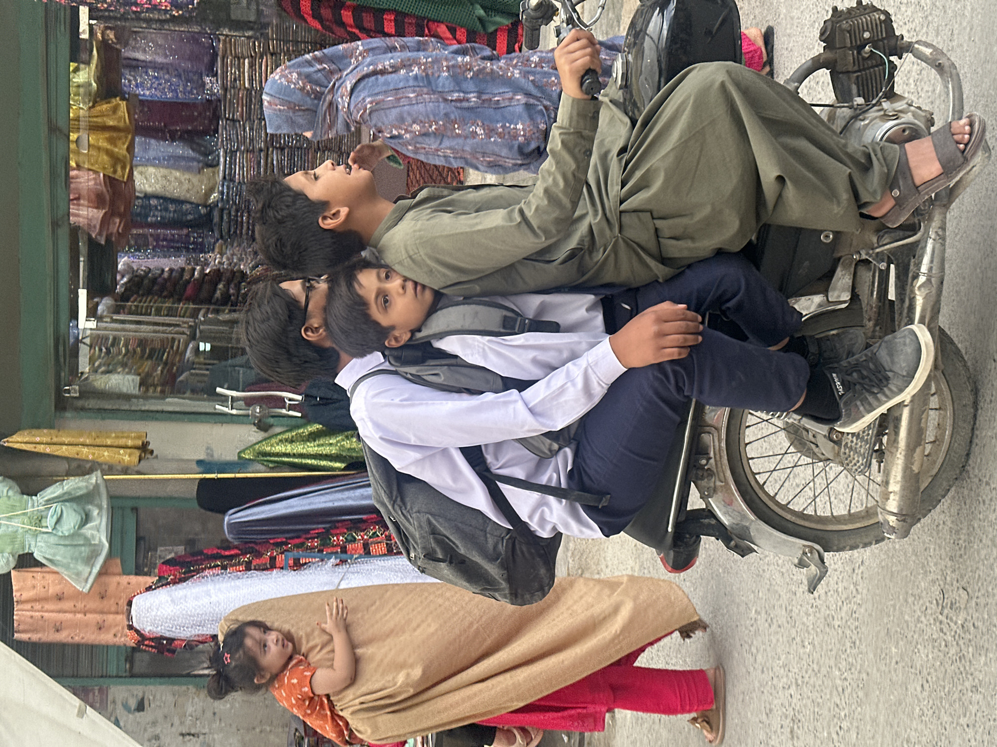 The image depicts three children on a motorcycle in Quetta City Tehsil, Pakistan. The motorcycle is parked in front of a shop selling textiles. An older boy is sitting on the motorcycle seat with his hands on the handlebars. Another boy is sitting in front of him, resting his head on the older boy's back. A third, younger child is positioned in front of the second boy, wearing a school uniform and backpack. A young girl is walking in front of the motorcycle wearing a orange shirt, tan shalwar kameez and pink pants. The background shows the shop with a variety of fabrics.
