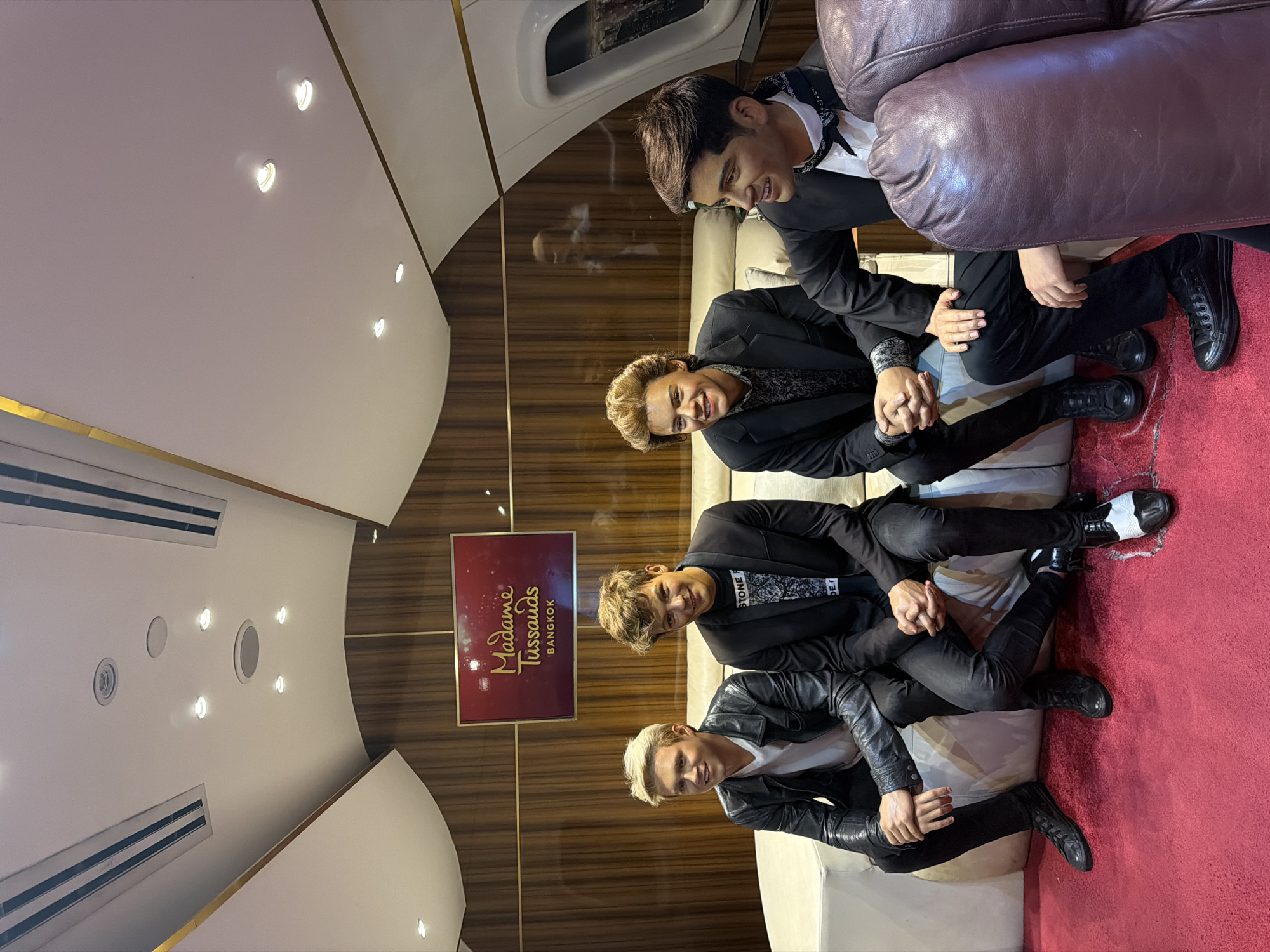 The image depicts a scene inside Madame Tussauds, Bangkok, Thailand. Four male wax figures, resembling the members of the band One Direction, are seated on a cream-colored couch. The setting appears to be designed to simulate the interior of a private jet, with wood-paneled walls, a curved ceiling with recessed lighting, and a window on the right side showing a blurry exterior. A red carpet covers the floor. The wax figures are dressed in fashionable attire. From left to right, the first figure wears a white t-shirt and a black leather jacket. The second figure is in a black suit jacket over a patterned shirt. The third figure is in a black suit jacket over a printed shirt. The fourth figure is in a black suit jacket with a white shirt and a black and white patterned bandana tied around his neck. The figures are posed as if interacting with each other, with some looking at each other and others looking forward. A framed Madame Tussauds Bangkok sign is visible on the wood-paneled wall behind the figures.