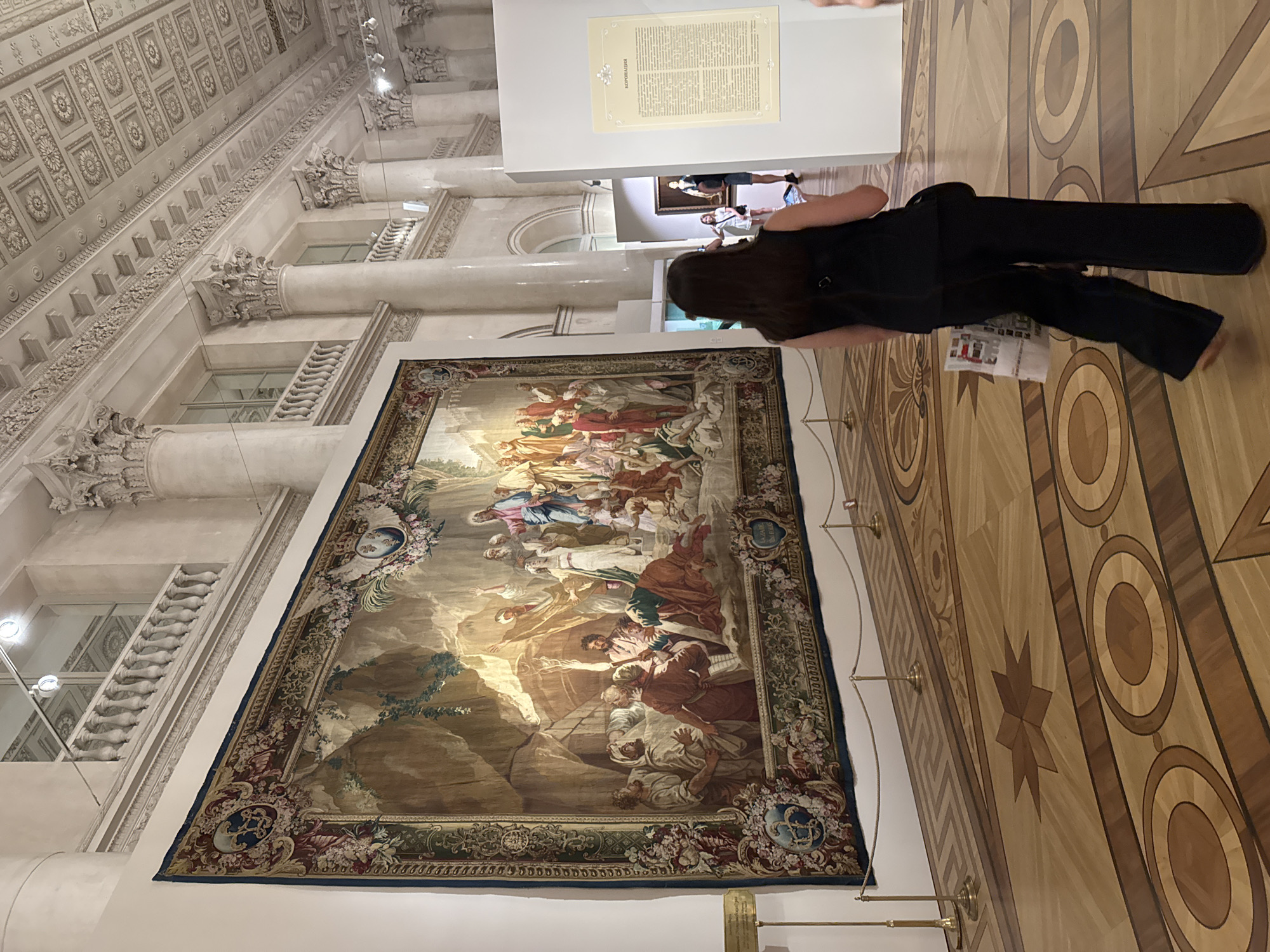 The image shows the interior of the Winter Palace in Saint Petersburg, Russia, specifically within the Hermitage Museum. A large, intricate tapestry depicting a biblical scene, possibly the Raising of Lazarus, hangs on the left wall. The tapestry features a complex composition with numerous figures dressed in ancient attire, set against a dramatic rocky landscape with shafts of light. The room itself is grand, with tall Corinthian columns, ornate ceiling decorations, and a highly polished parquet floor with geometric and circular inlays.
In the foreground, a woman with long dark hair, wearing a black sleeveless top and dark wide-leg pants, is walking away from the camera, looking at a phone in her right hand and holding a folded brochure or pamphlet in her left. She is barefoot. Behind her, a white display stand with text in Russian is visible, with the title "КОРОНАЦИЯ" (Coronation) prominently displayed. Further in the background, other museum visitors and artworks can be seen, including a painting on an easel. The overall impression is one of opulence and historical significance, typical of the museum's galleries.