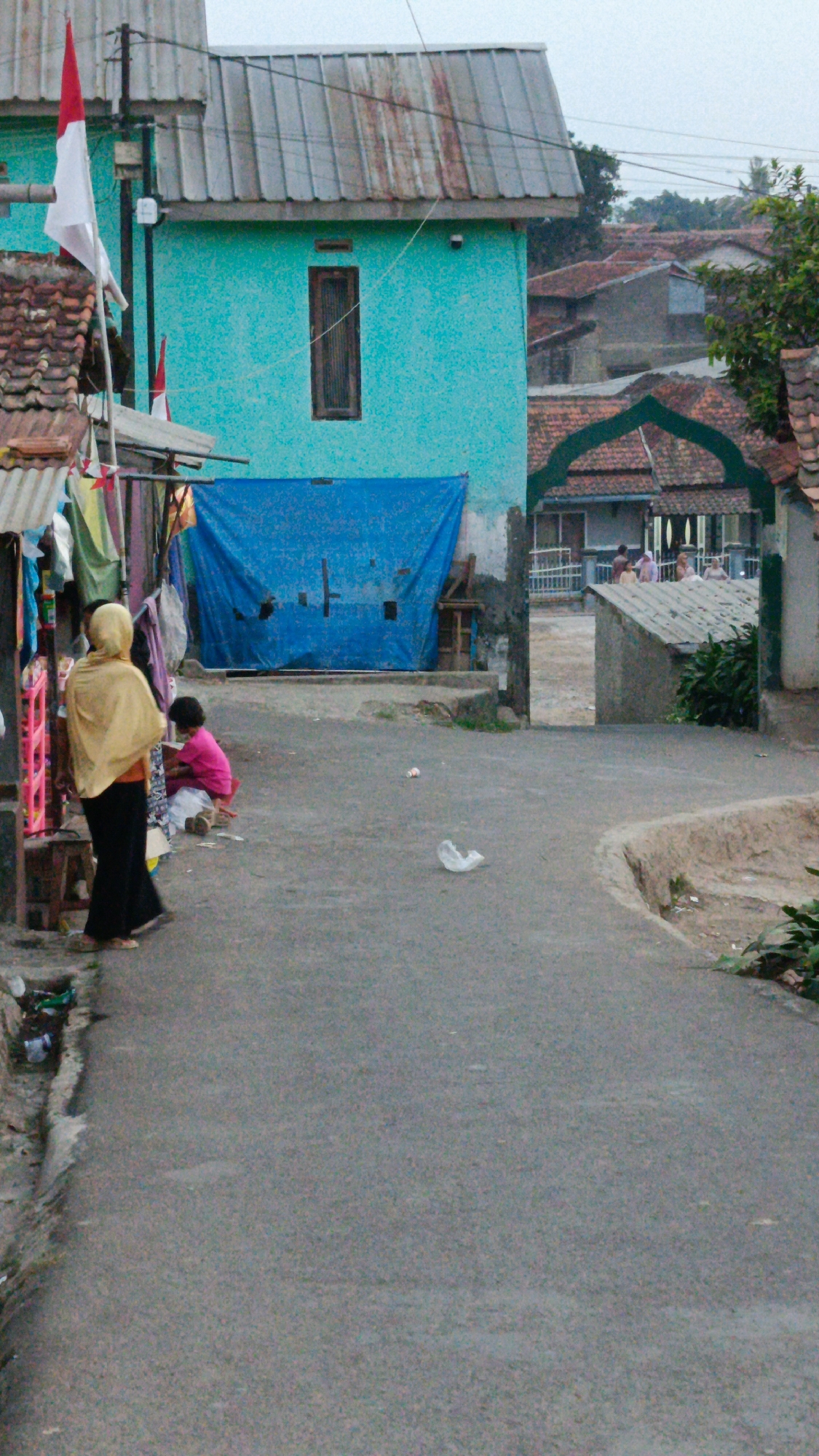 In Margajaya, Indonesia, a street scene unfolds with a bright turquoise building dominating the left side. A corrugated metal roof, showing signs of rust, covers the structure. A dark wooden-framed window is visible on the turquoise facade. To the left of the building, a smaller structure with a similar rusted metal roof has a partially visible white and red flag attached to a pole. A blue tarp is draped over an opening beneath the turquoise building.
In the foreground, a woman wearing a yellow headscarf and dark pants stands facing away from the camera. She is positioned near a stall or shop displaying various items of clothing. To her right, a young child, wearing a bright pink shirt and white pants, is seated on the ground, partially obscured. The street itself is paved with asphalt and appears somewhat worn. Scattered plastic debris is visible on the street.
Beyond the turquoise building, a row of houses with red tile roofs extends into the background. An arched gateway with a red-tiled roof frames a view of more houses and a few people standing in the distance. Green foliage is present in the upper right portion of the image. The overall impression is of a modest, lived-in urban environment.