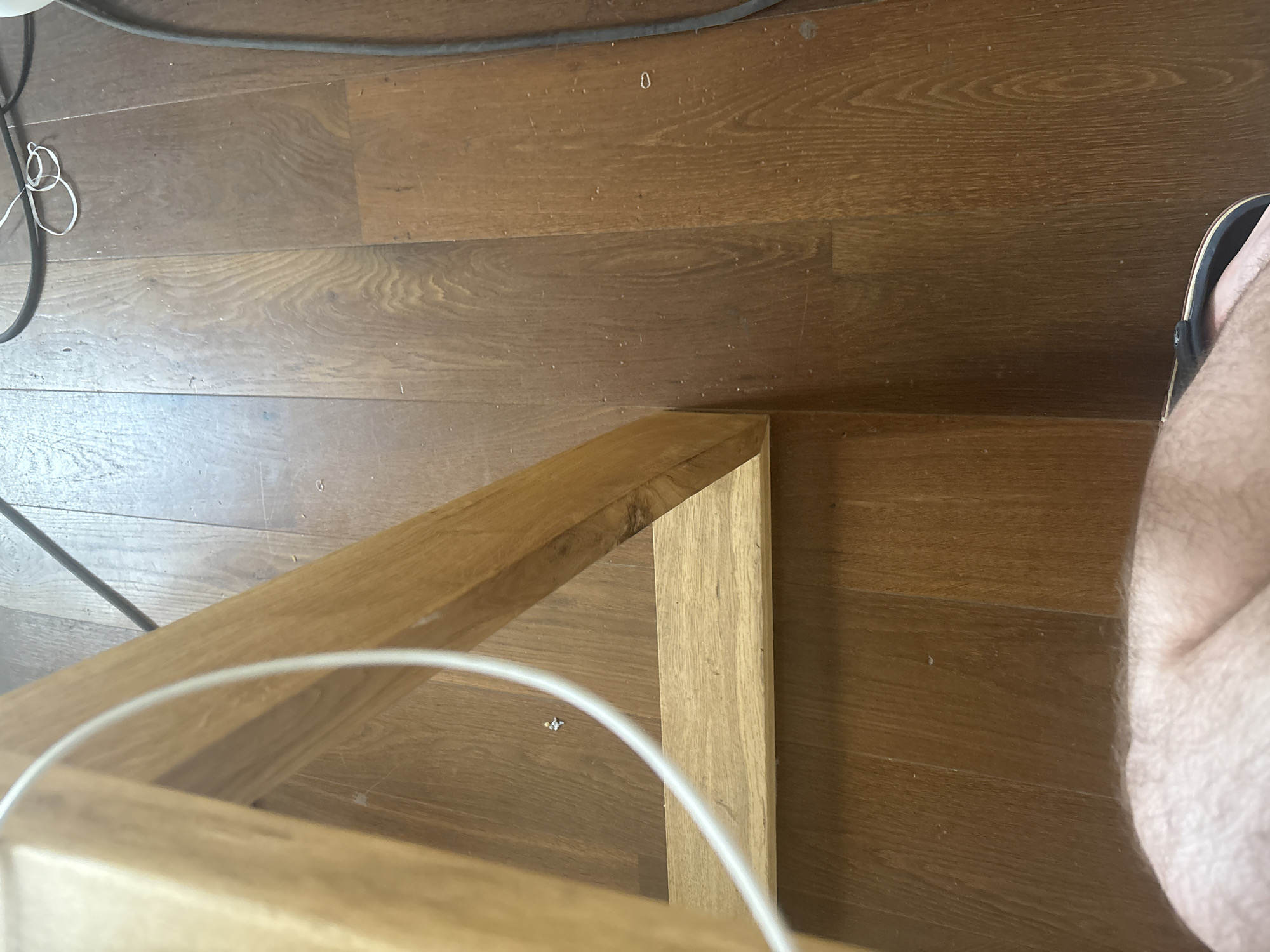 The image shows the floor of a room, likely a home. The floor is made of wooden planks, with a noticeable grain pattern. A portion of a wooden table leg and a white cable are visible on the left side of the frame. In the background, a black cable is seen running along the floor. The lower part of the image shows a person's leg and foot wearing a sandal. The scene is domestic and appears to be an indoor setting. There is no identifiable information within the image that suggests a specific location in the city of Ореховка, Russia.