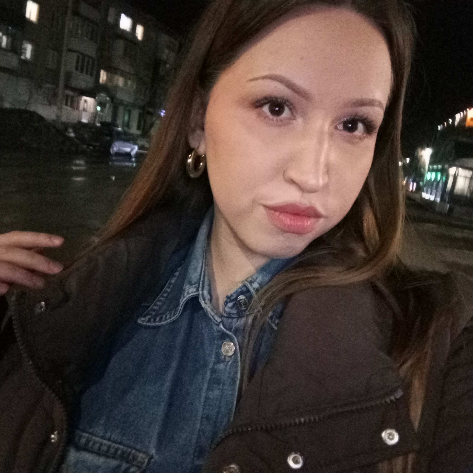 A young woman is taking a selfie outdoors at night. She has long brown hair, light skin, and is wearing large hoop earrings. She is dressed in a blue denim shirt under a dark jacket. In the background, there are buildings with lit windows and streetlights. There appears to be a wet, reflective surface, suggesting rain. The general location is likely a street in the city of Chaikovsky, Russia, based on the prompt instructions, but there are no specific identifiers in the image to confirm the exact location.
